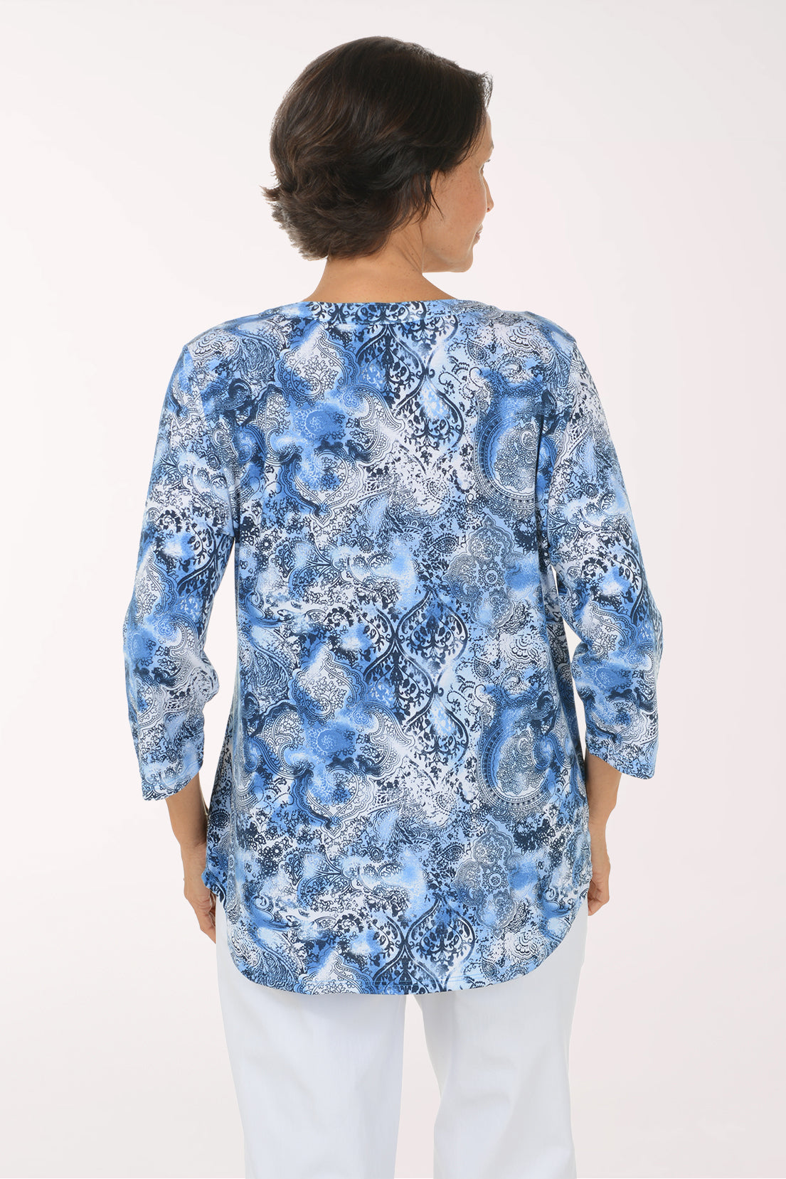 A woman modeling a blue and white abstract patterned long-sleeve top with a split neckline, paired with white pants, against a plain white studio background.