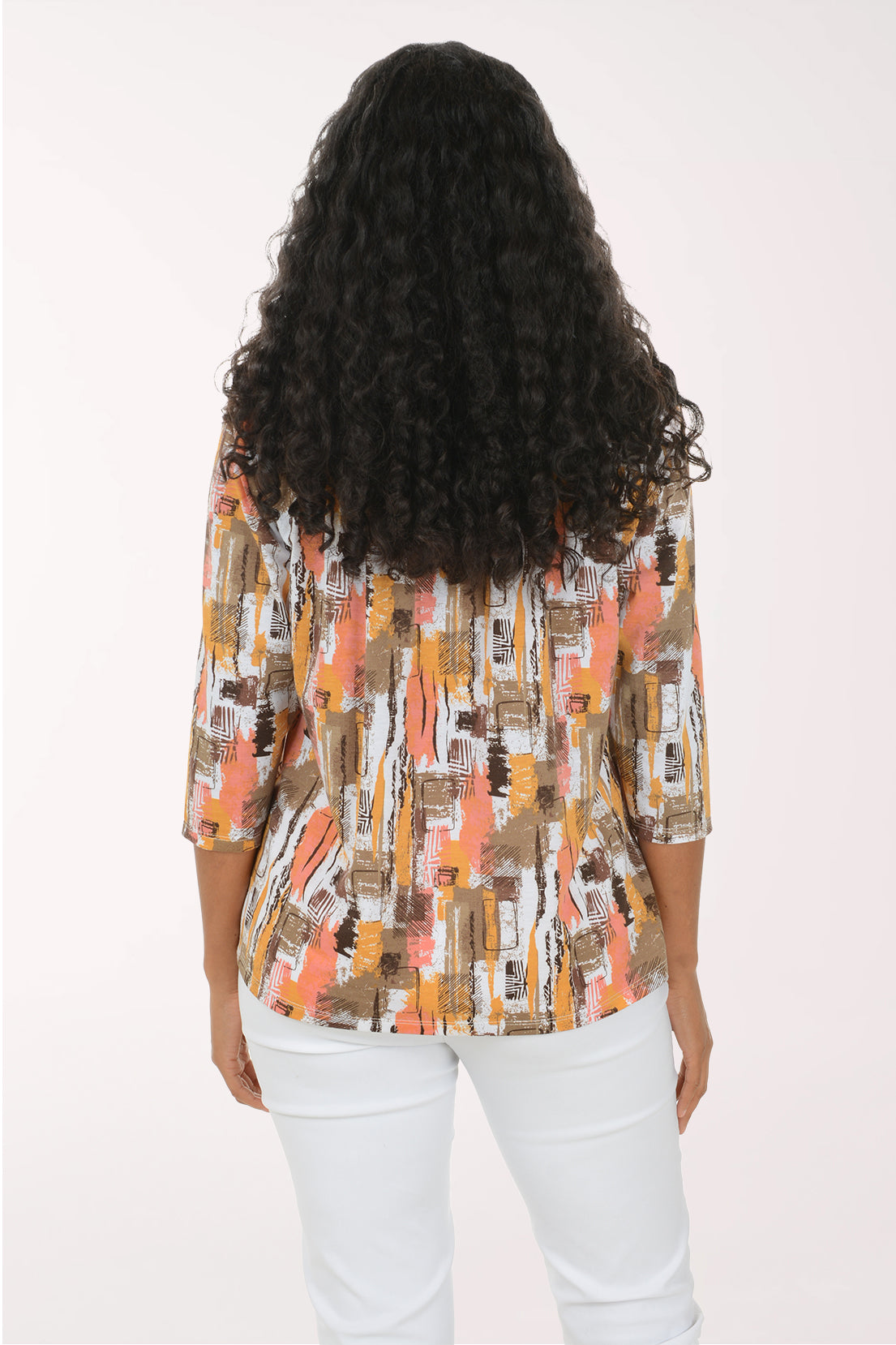 A smiling woman with long curly black hair models a three-quarter sleeve blouse with an abstract brushstroke pattern in orange, brown, beige, and white, paired with white pants against a plain white studio background.