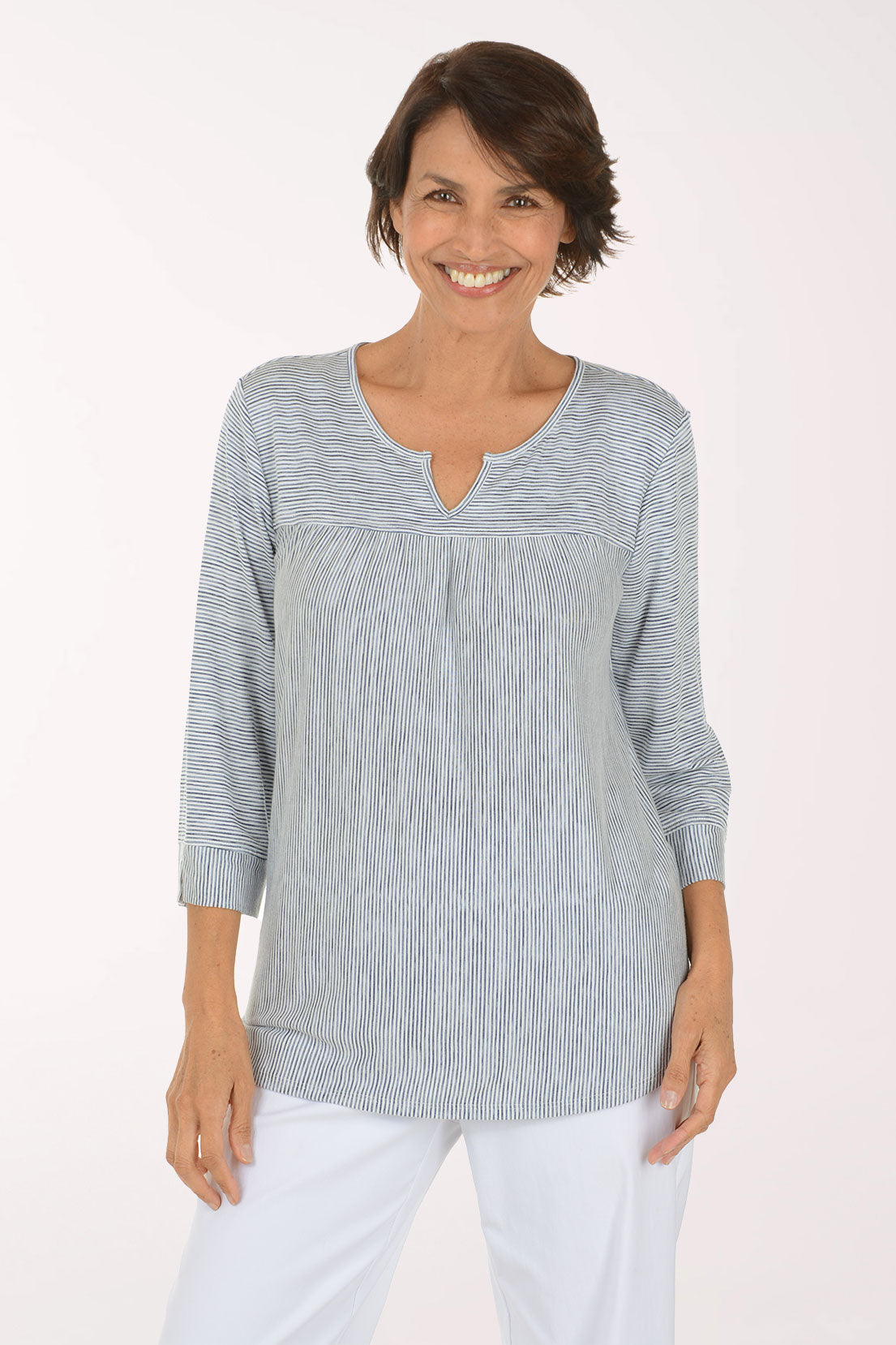 A smiling woman with short brown hair stands with arms crossed, wearing a light gray and white vertically textured long-sleeve top with a split neckline, paired with white pants, against a plain white studio background.