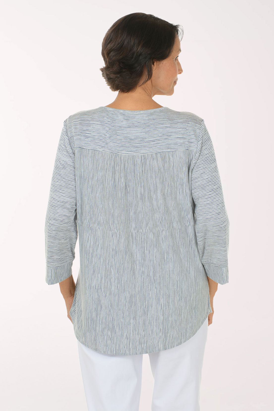 A smiling woman with short brown hair stands with arms crossed, wearing a light gray and white vertically textured long-sleeve top with a split neckline, paired with white pants, against a plain white studio background.