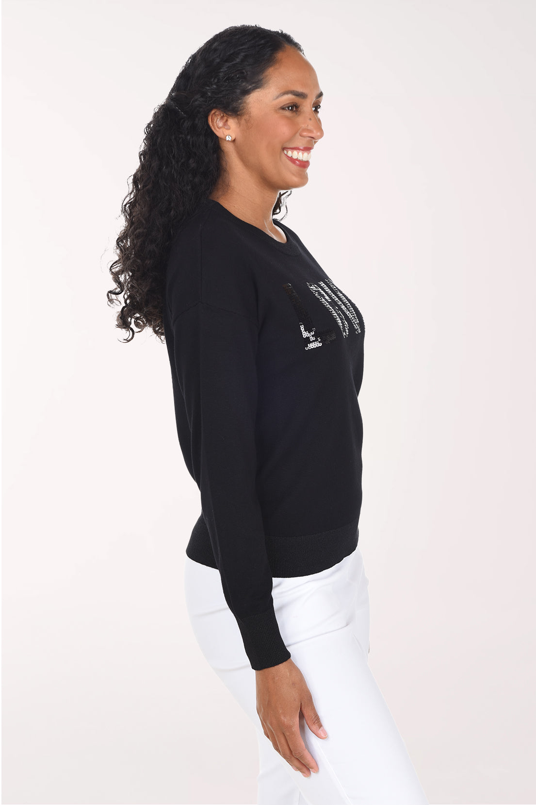 Side view of a woman wearing a black crewneck sweater with sequined "Love" lettering on the front, styled with white pants.