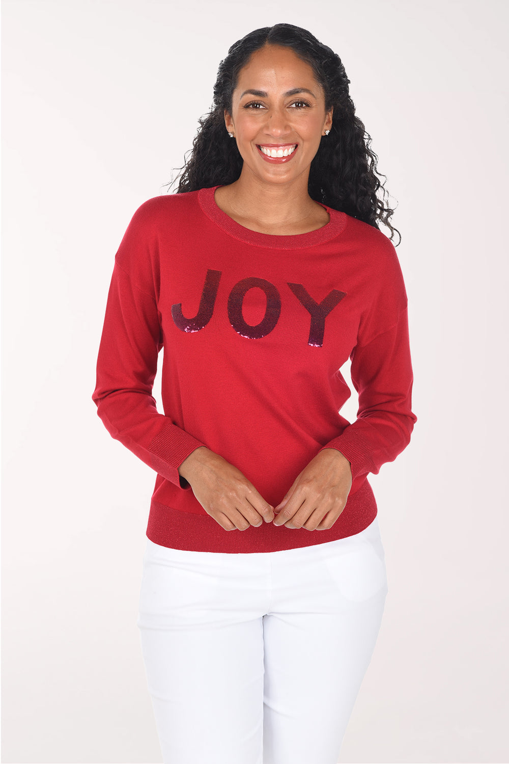 Front view of a woman wearing a red crewneck sweater with "JOY" in deep red sequined block letters, styled with white pants.