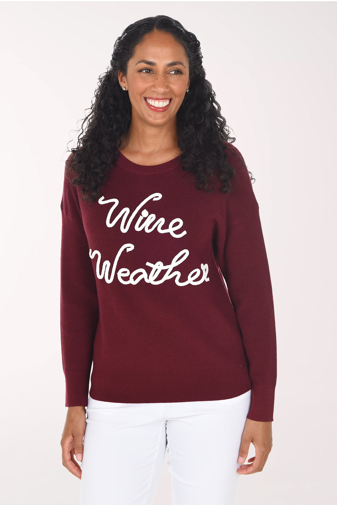 Front view of a woman wearing a deep wine red crewneck sweater with "Wine Weather" in white braided script across the front, styled with white pants.