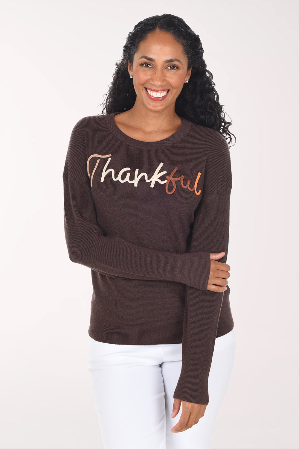 Front view of a woman wearing a chocolate brown crewneck sweater with "Thankful" in script, "Thank" in blush pink and "ful" in copper, styled with white pants.