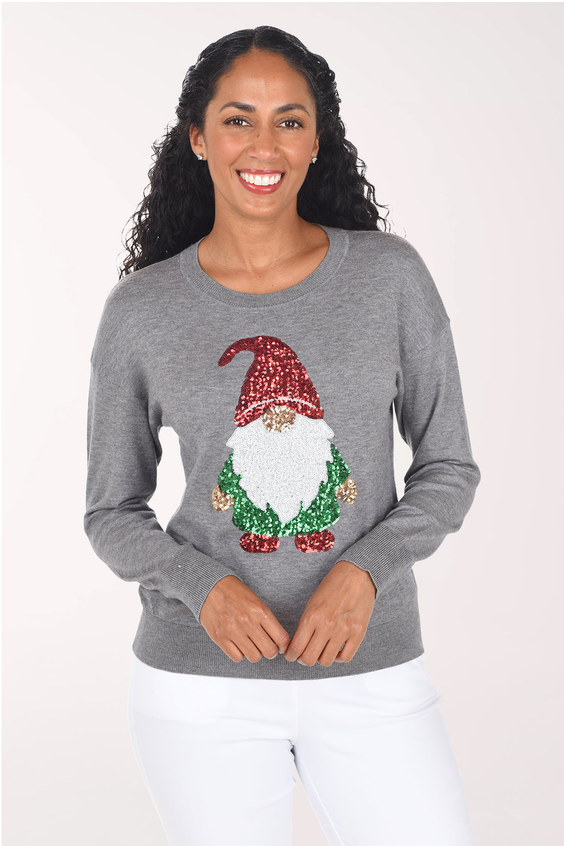 Front view of a woman wearing a heather grey crewneck sweater with a sequined holiday gnome on the front: red sequin hat, fuzzy white beard, gold nose and green sequin body, styled with white pants.