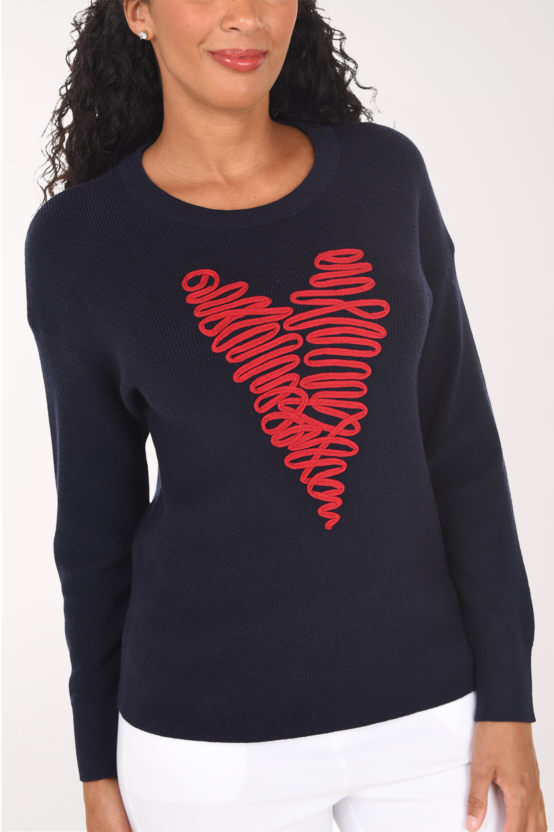 A woman wearing a navy blue crewneck sweater with a large red wavy graphic on the front, paired with white pants, against a plain light background.