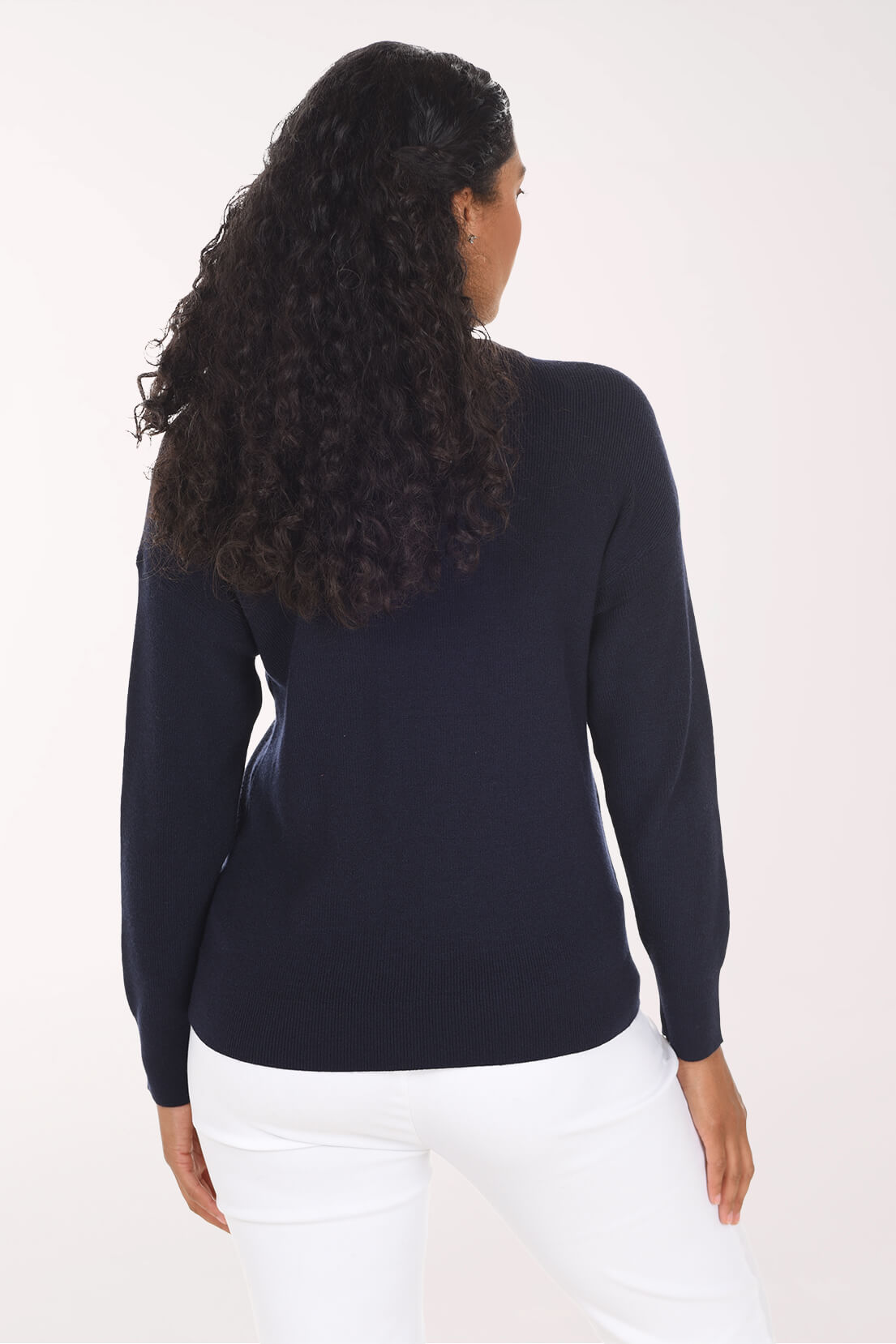 A woman with long, curly dark hair is shown from the back wearing a dark navy blue long-sleeve knit top and white pants against a plain light background.