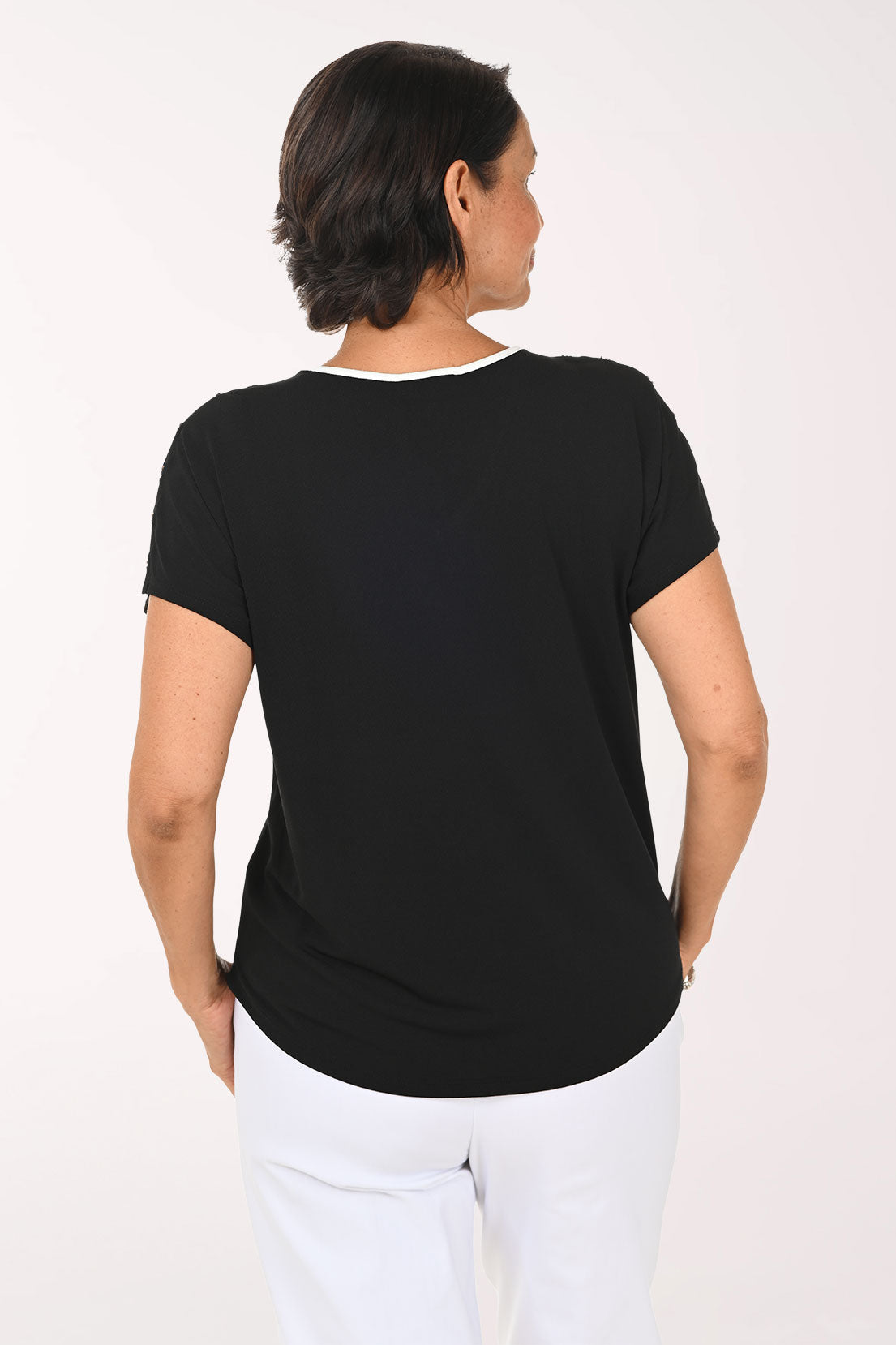 A woman shown from the back wearing a black short-sleeve t-shirt with a thin white inner collar trim and white pants, set against a plain white studio background.