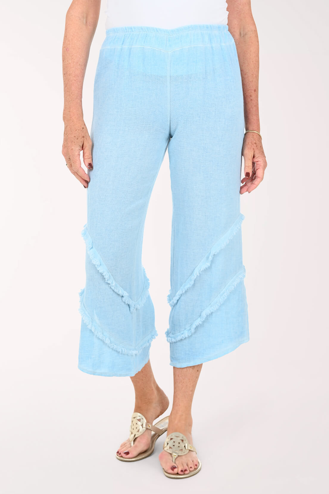 A person wearing light blue cropped pants with layered ruffle details near the hems, shown against a plain light background. The model is also wearing beige slide sandals.