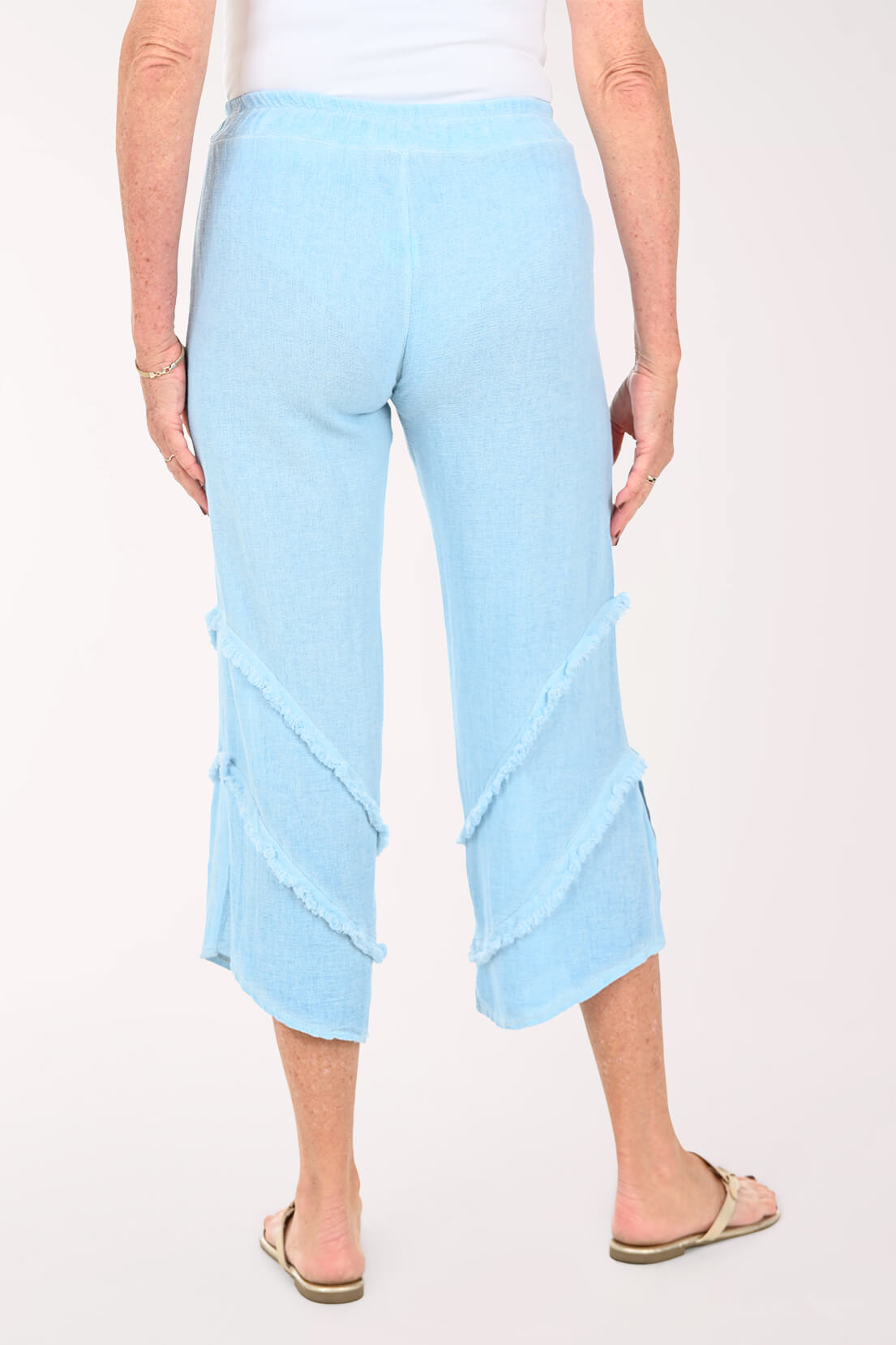 A person shown from the back wearing light blue cropped pants with diagonal ruffle details and side buttons, paired with a white top and tan sandals, against a plain light background.