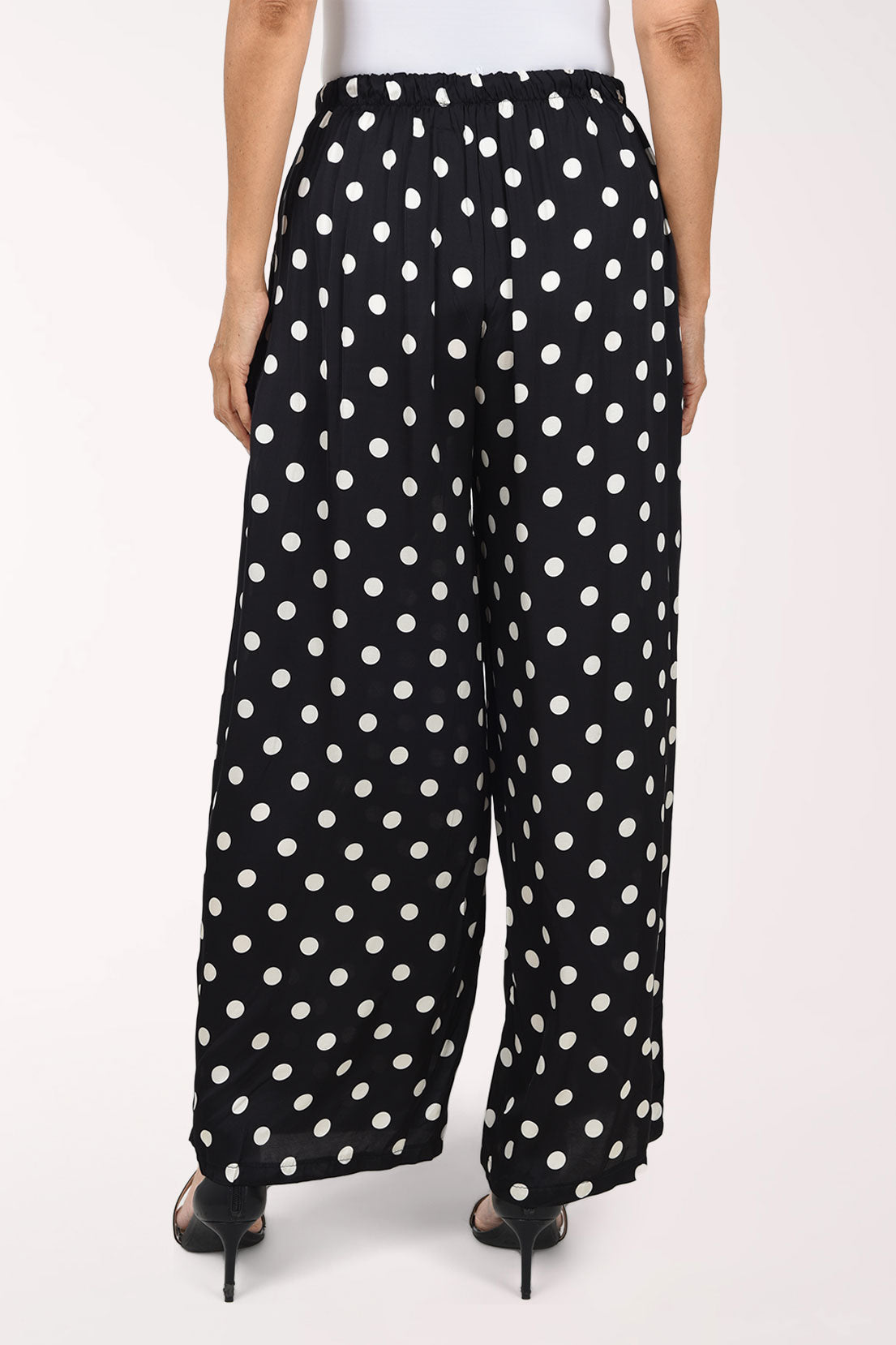 Black wide-leg pants with a white polka dot pattern, shown on a model wearing black strappy heels against a plain white studio background.