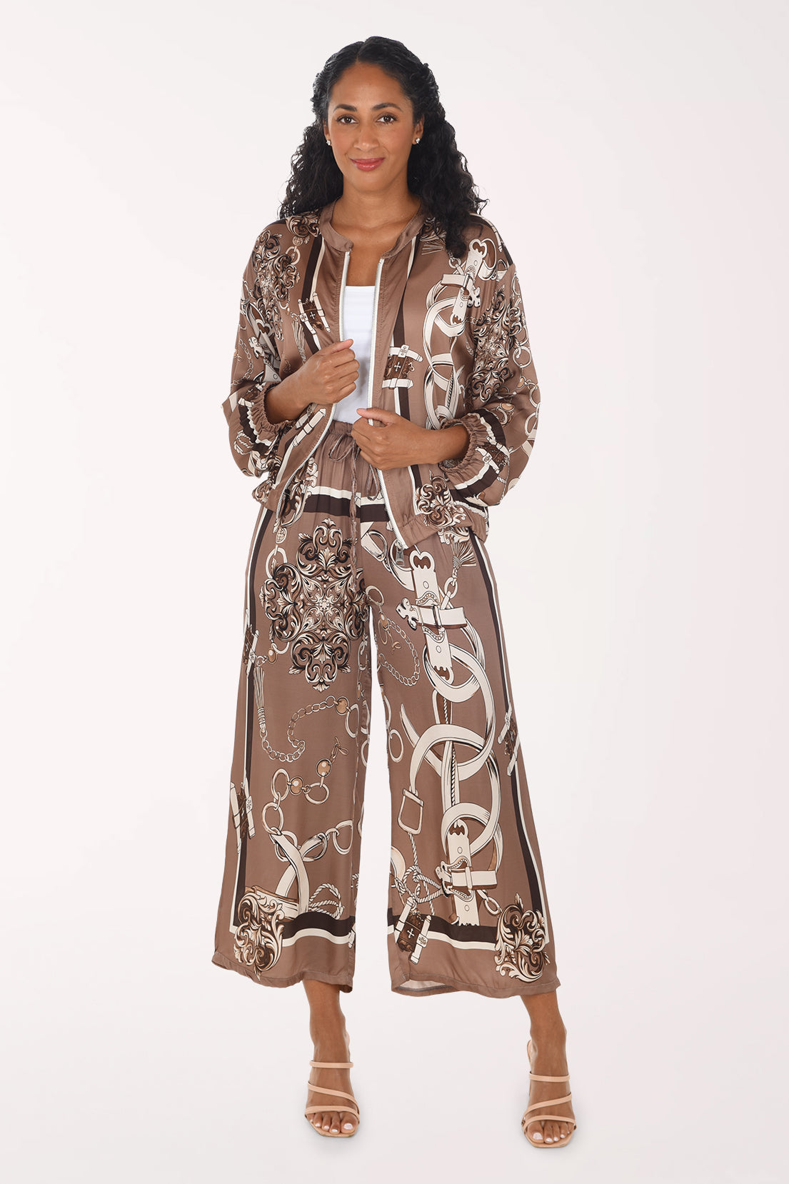 A woman modeling a matching two-piece set in a brown and beige silk-like fabric with a white graphic print. The set includes a cropped jacket with a drawstring and wide-leg pants, styled with a white top and beige strappy heels against a plain white background.