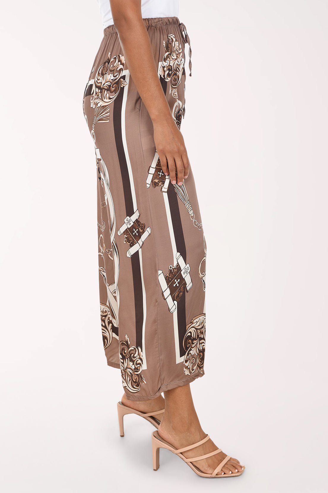 A side-profile view of a model wearing brown wide-leg cropped pants with a black, white, and beige graphic print, paired with beige high-heeled sandals against a light off-white background.