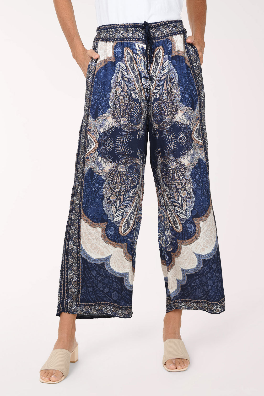 A model wearing wide-leg cropped pants with a detailed blue, beige, and white paisley and floral pattern, paired with beige slide sandals against a plain light background.