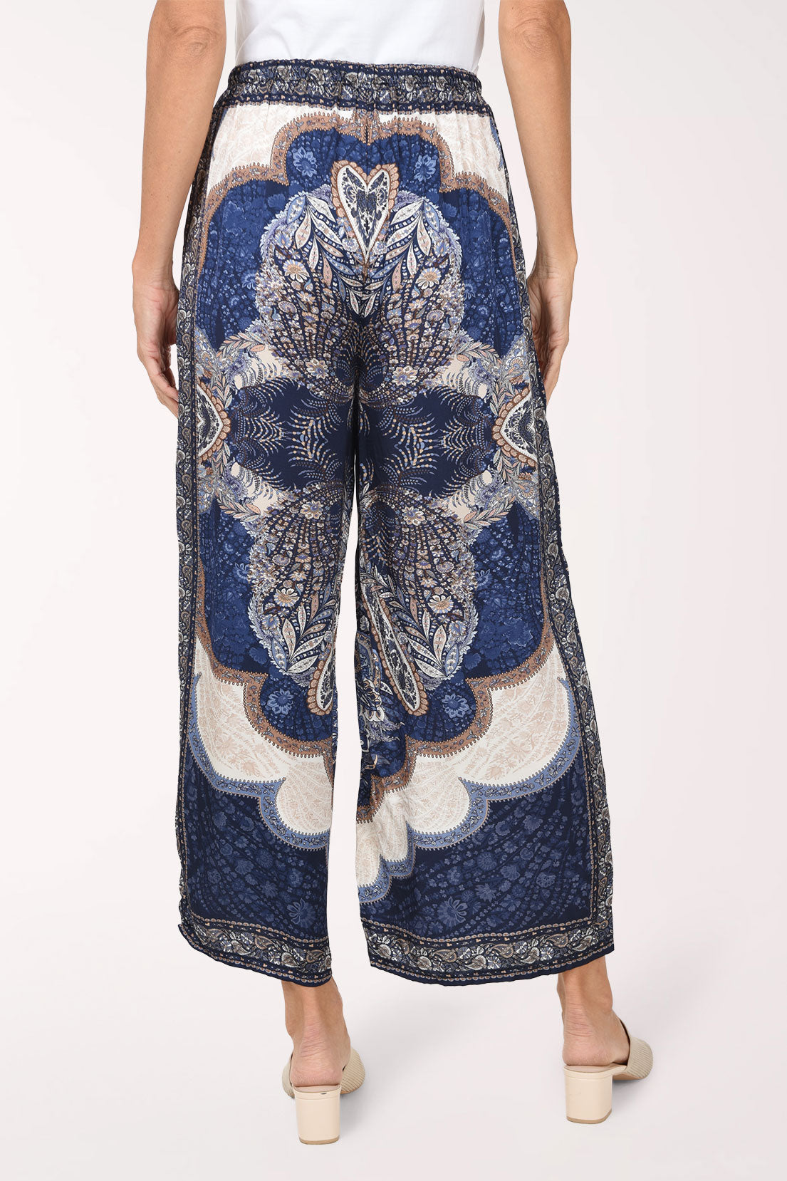 A person shown from the back wearing wide-leg cropped pants with a blue, white, and beige ornate medallion pattern, paired with a white top and beige heeled sandals against a light studio background.