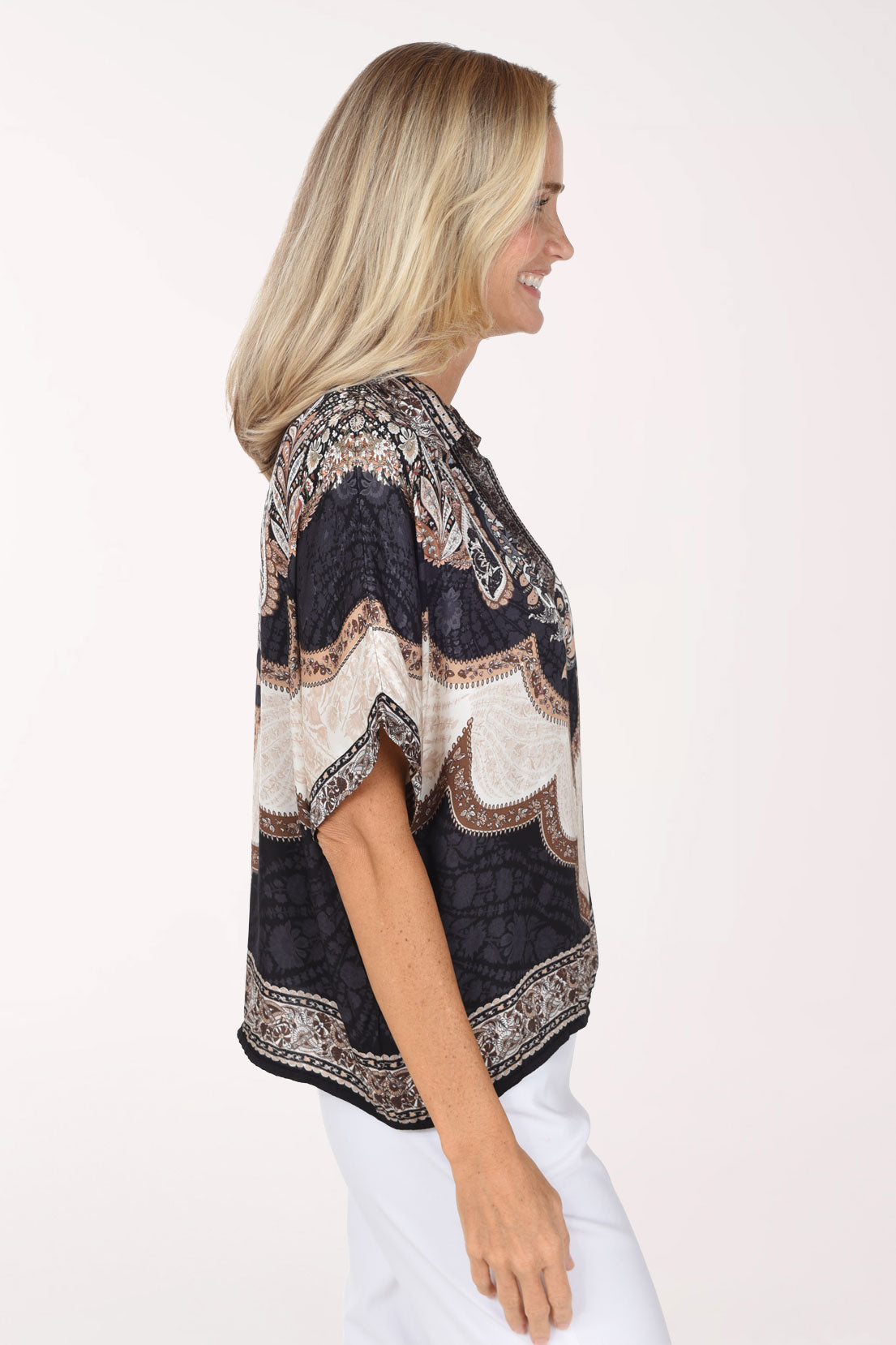 A smiling blonde woman wearing a short-sleeve V-neck top with a black, beige, and brown paisley pattern, paired with white pants, against a plain white studio background.