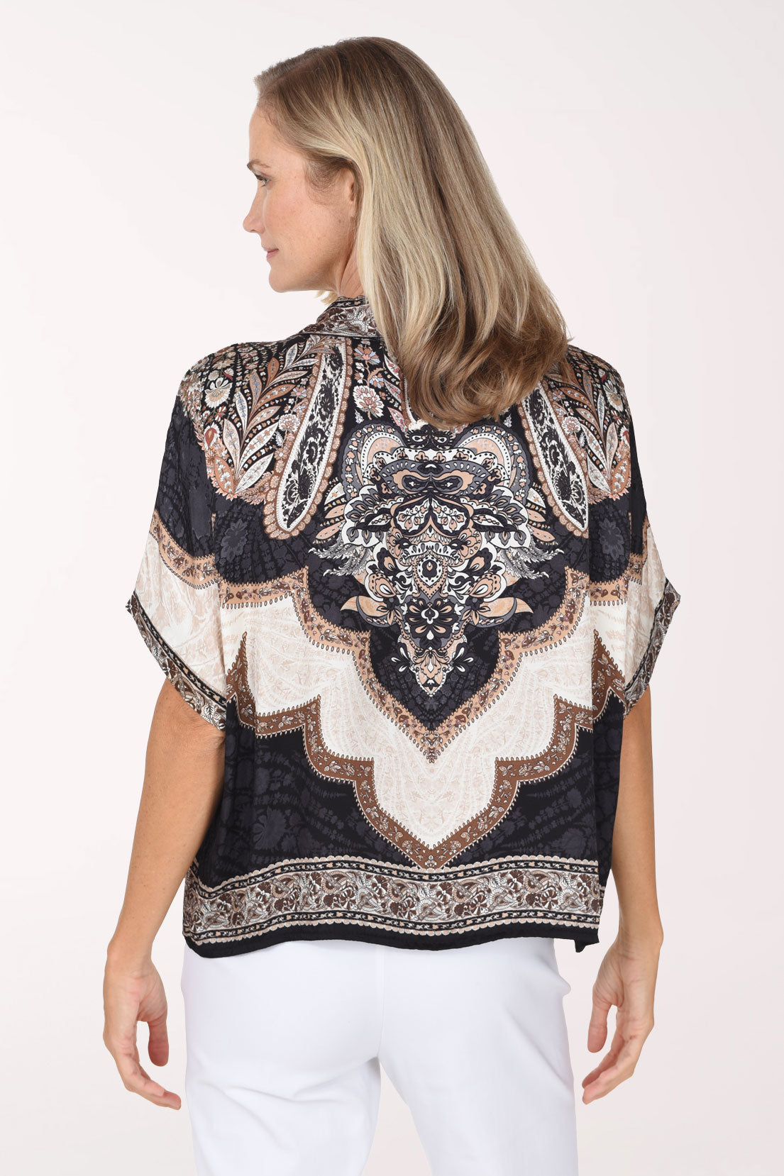 A smiling blonde woman wearing a short-sleeve V-neck top with a black, beige, and brown paisley pattern, paired with white pants, against a plain white studio background.