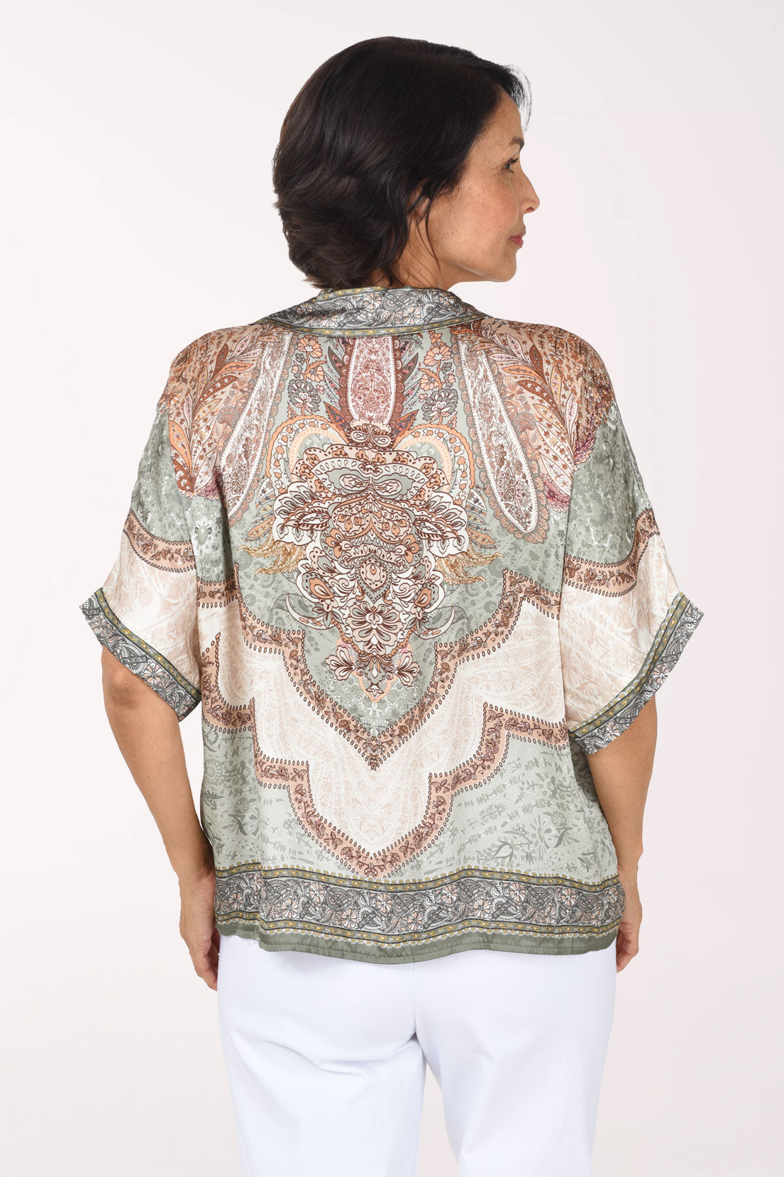 A smiling woman wearing a short-sleeve V-neck blouse with an intricate paisley and floral pattern in beige, sage green, and brown, paired with white pants, against a plain light background.