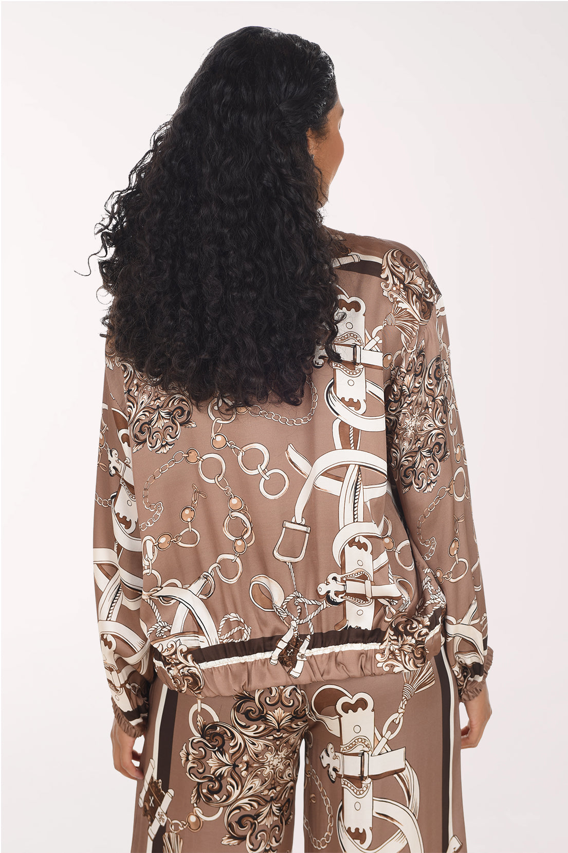 A woman modeling a brown satin zip-up jacket and matching pants set with a white and brown baroque-style pattern, worn over a white tank top, against a plain light background.