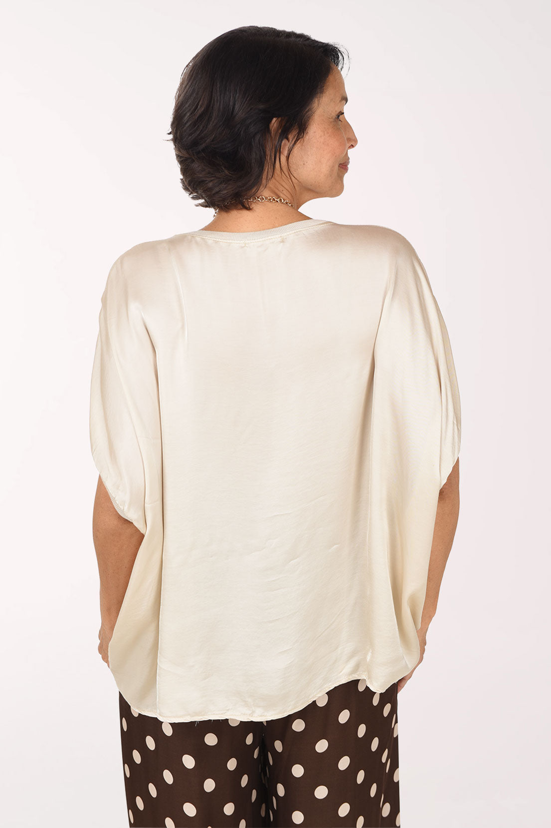A smiling woman wearing a cream-colored short-sleeve V-neck top with a silky sheen, paired with brown and white polka dot pants and a long gold-toned necklace, set against a plain white studio background.
