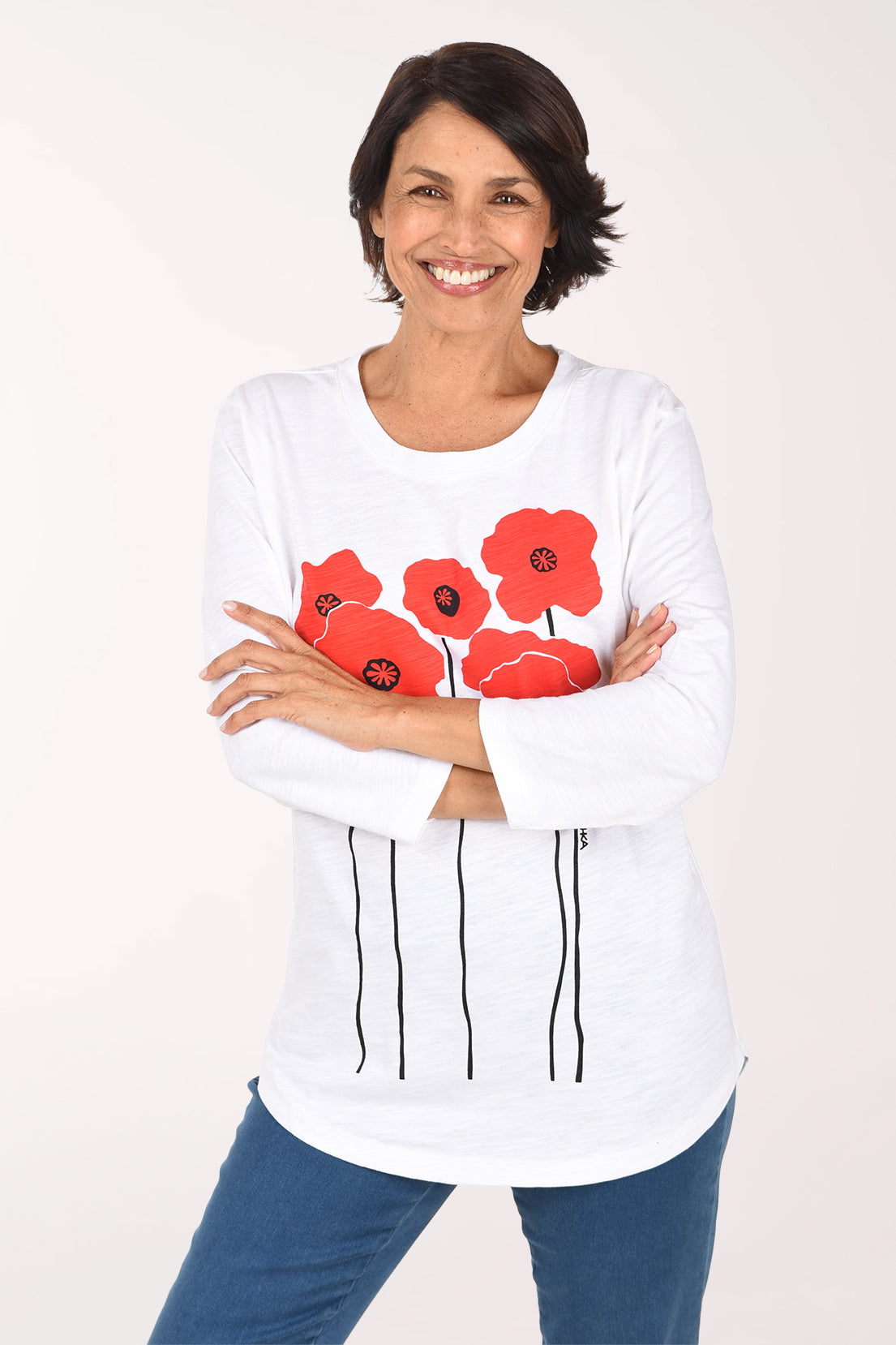 Smiling woman with arms crossed in a white 3/4 sleeve tunic top with a front graphic of bold red poppies on tall black stems, worn with blue jeans