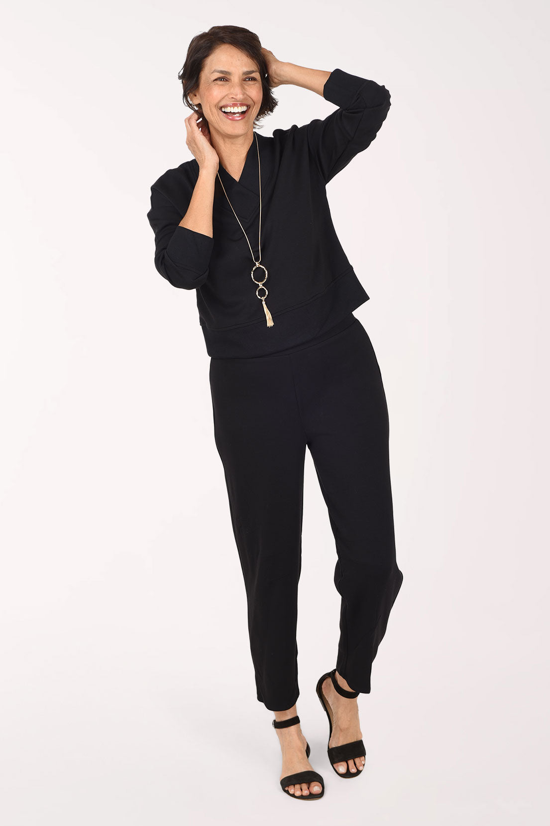 A woman wearing a black long-sleeve top and matching black pants, standing against a plain white studio background.