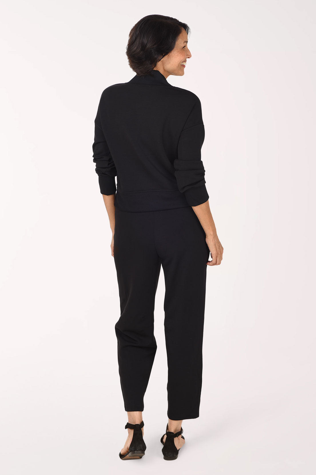 A woman wearing a black long-sleeve top and matching black pants, standing against a plain white studio background.