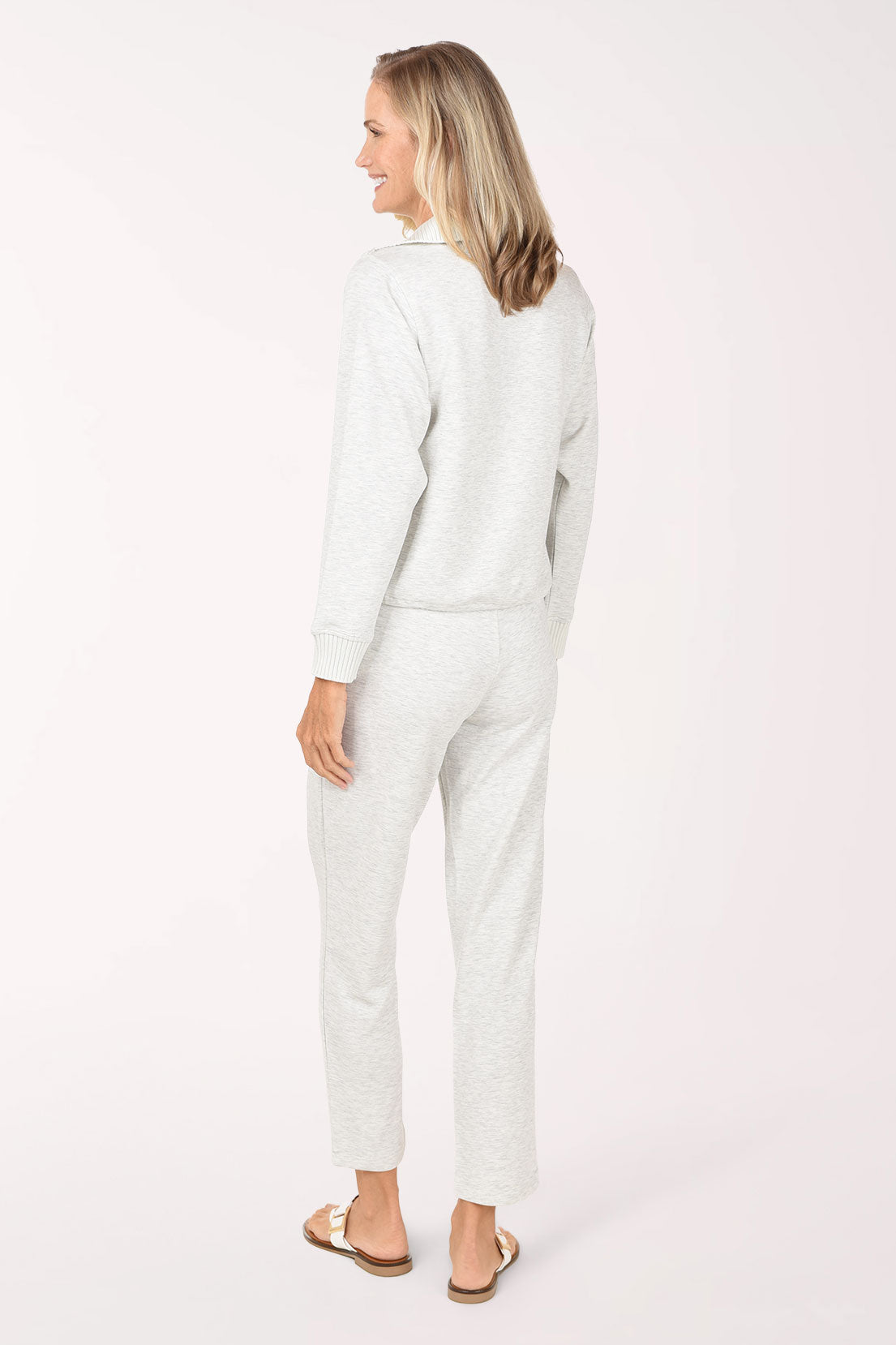 A woman wearing a light grey half-zip sweater and matching grey pants, standing against a plain white studio background.