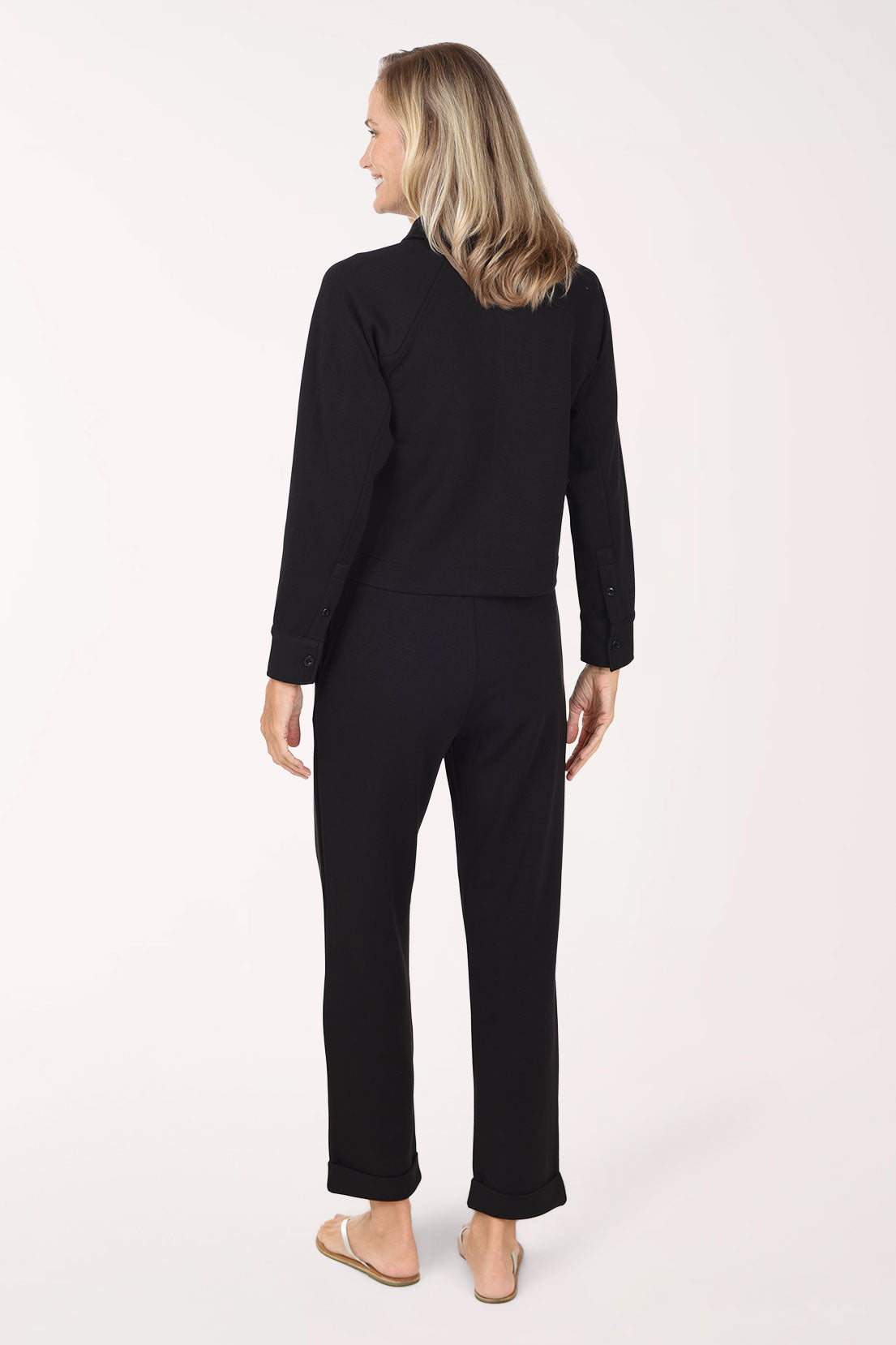 A woman wearing a black long-sleeve top and matching black pants, standing against a plain white studio background.