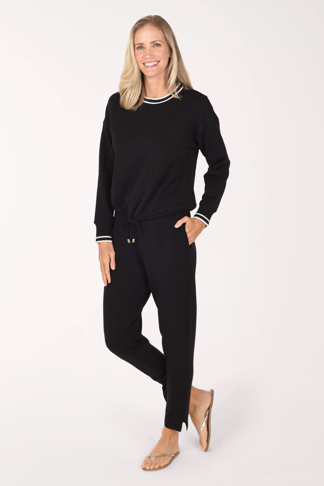 A woman modeling a black two-piece lounge set consisting of a long-sleeve crewneck top with white striped trim and matching black drawstring pants, set against a plain white studio background.