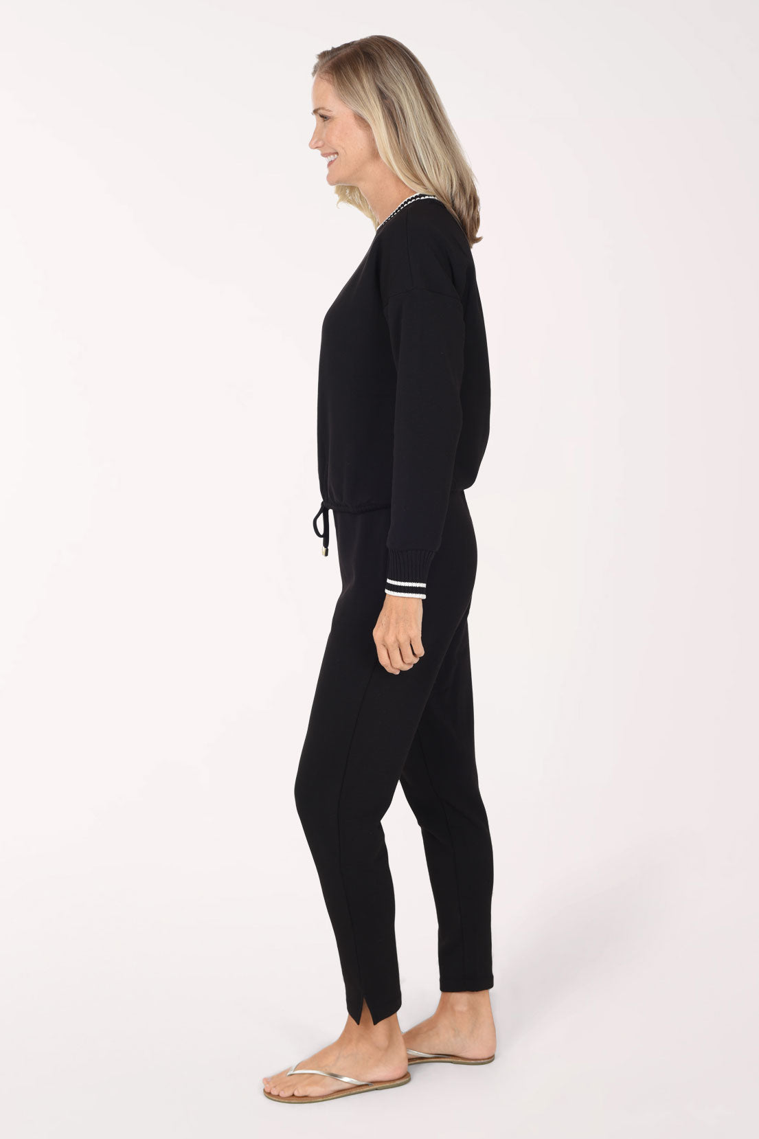 A woman in a black long-sleeve knit top and matching black pants stands in profile against a plain white studio background, wearing light-colored flip-flop sandals.