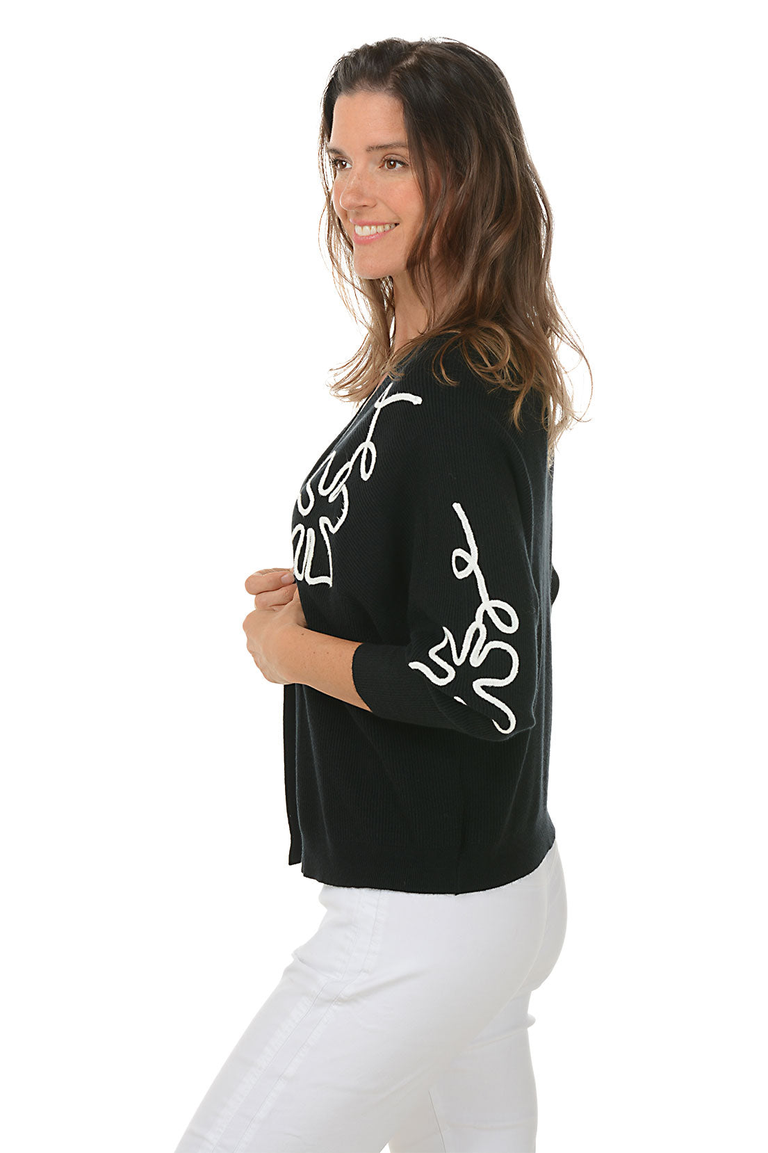 Woman wearing a black cardigan with white floral patterns on a white background. Side shot.