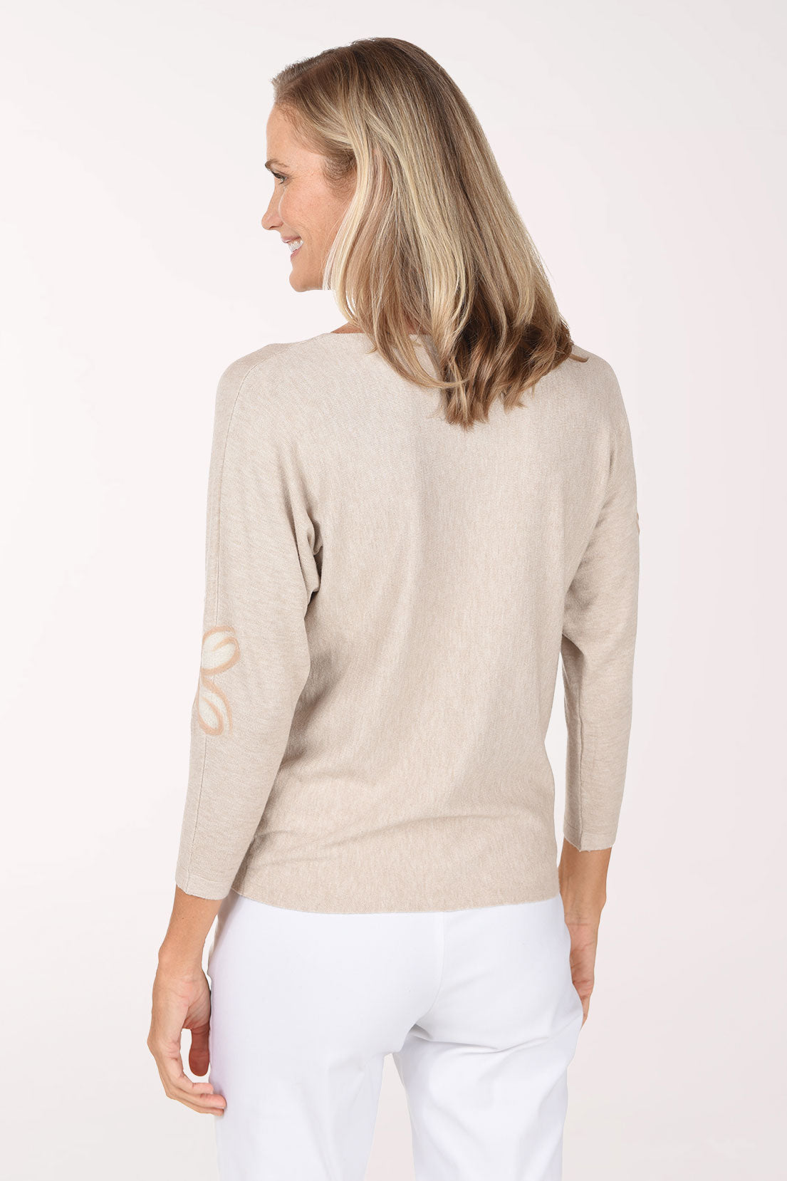 A woman wearing a light beige knit sweater with a tonal embroidered flower details, paired with white pants, against a plain white studio background.