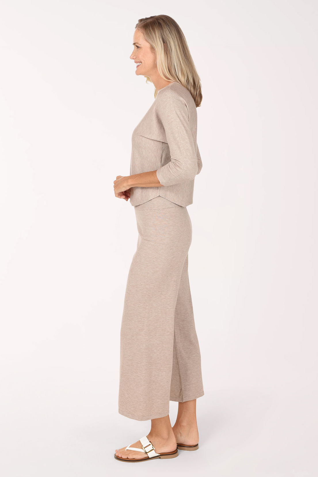 A woman modeling a matching two-piece knit loungewear set in a light taupe-beige color, featuring a three-quarter sleeve top and wide-leg cropped pants, paired with white slide sandals against a plain white studio background.