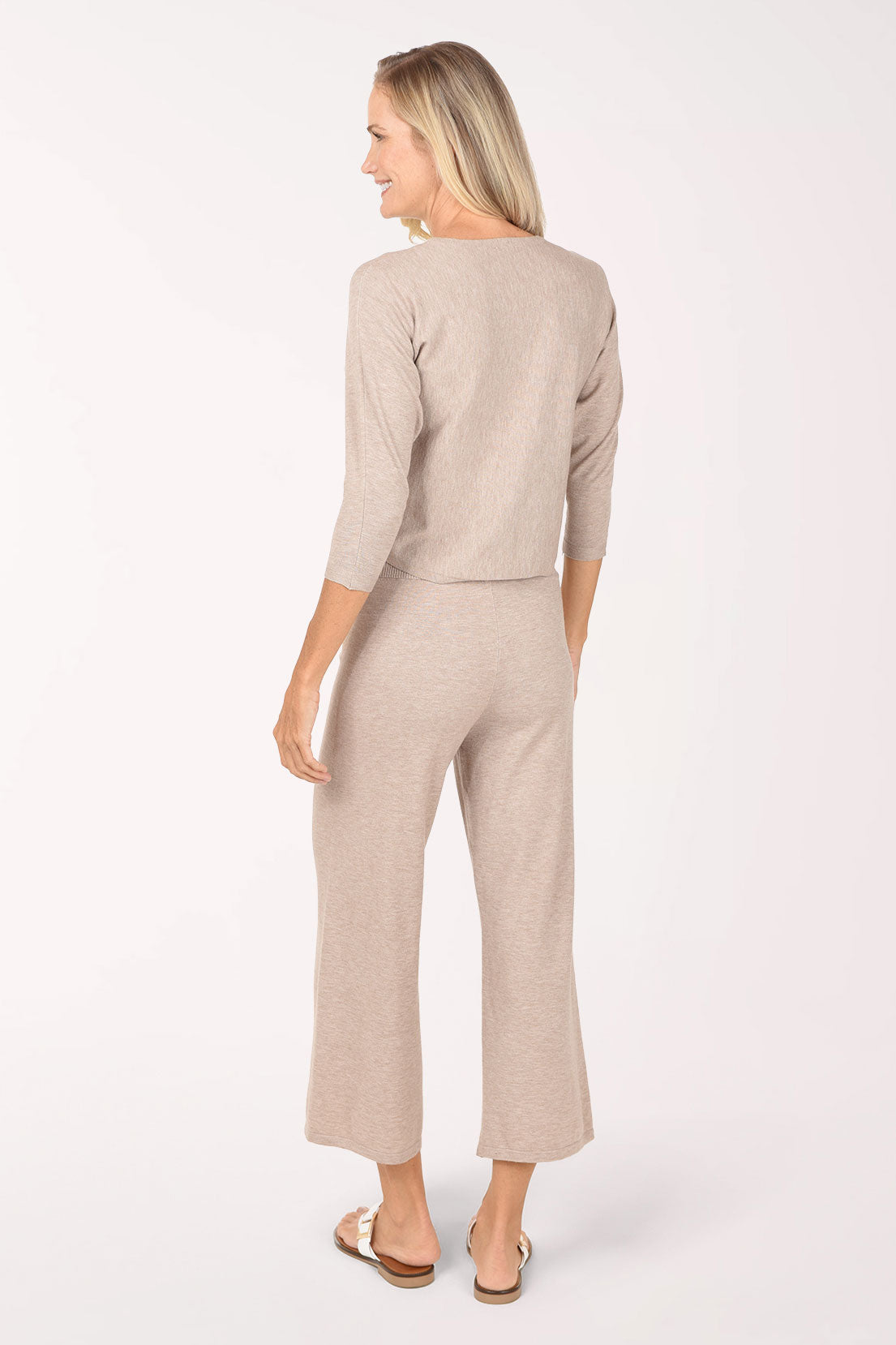 A woman modeling a matching two-piece knit loungewear set in a light taupe-beige color, featuring a three-quarter sleeve top and wide-leg cropped pants, paired with white slide sandals against a plain white studio background.