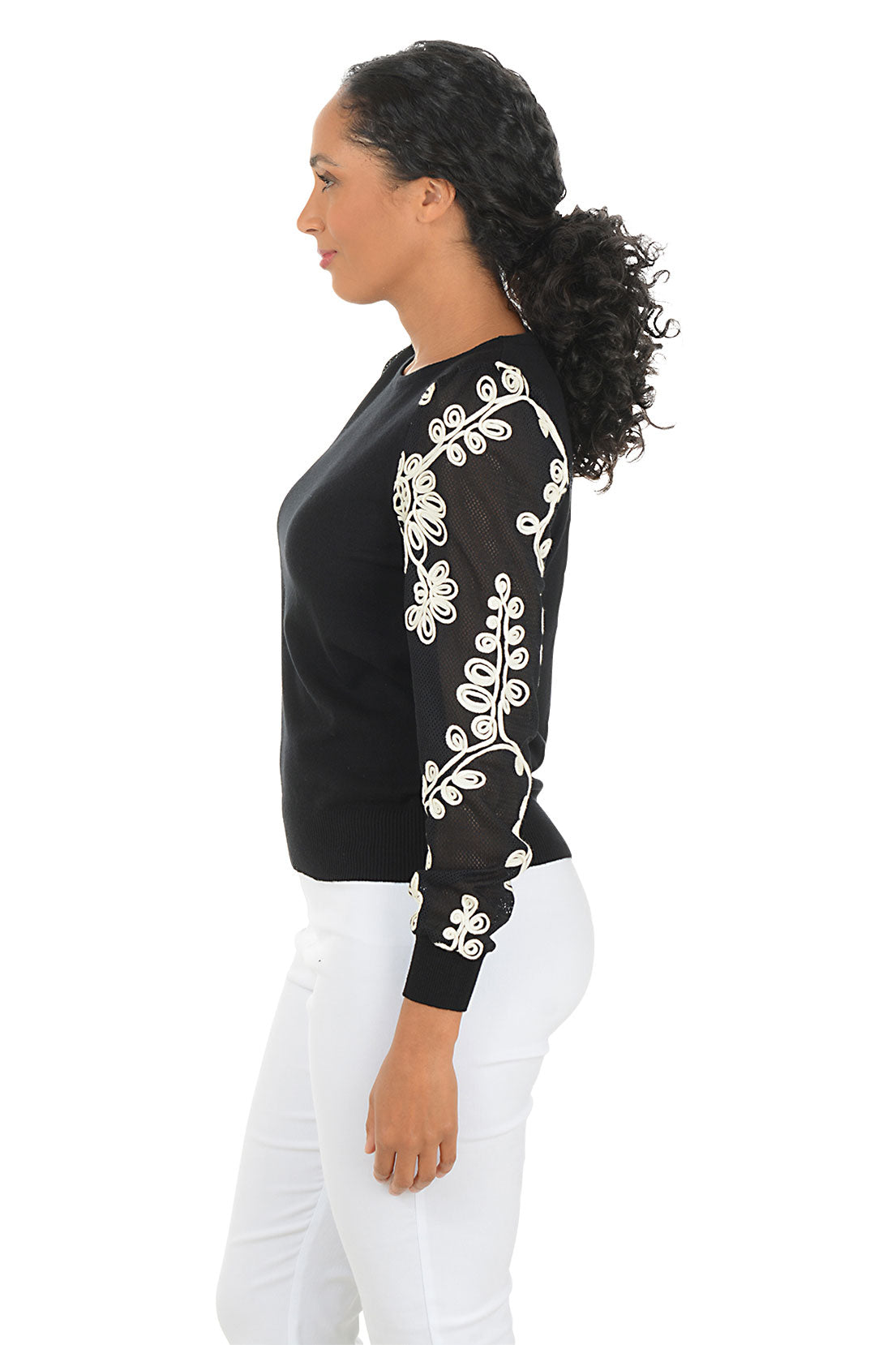 Woman wearing a black top with white embroidery on sleeves against a white background. Side shot.