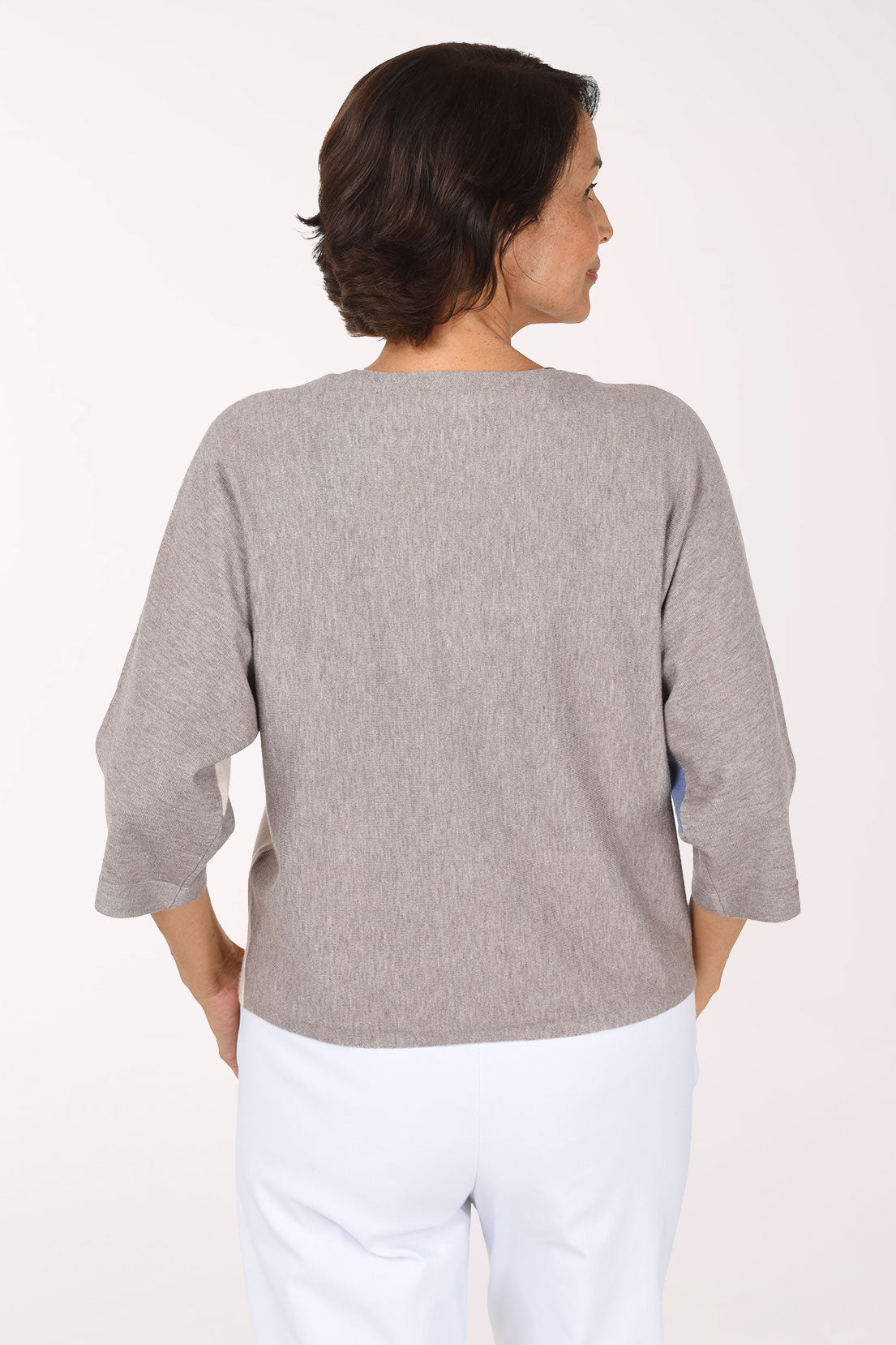 A woman modeling a color-block knit sweater with three-quarter sleeves, featuring a heather gray body, a light blue front panel, and a cream side panel, paired with white pants against a plain white studio background.
