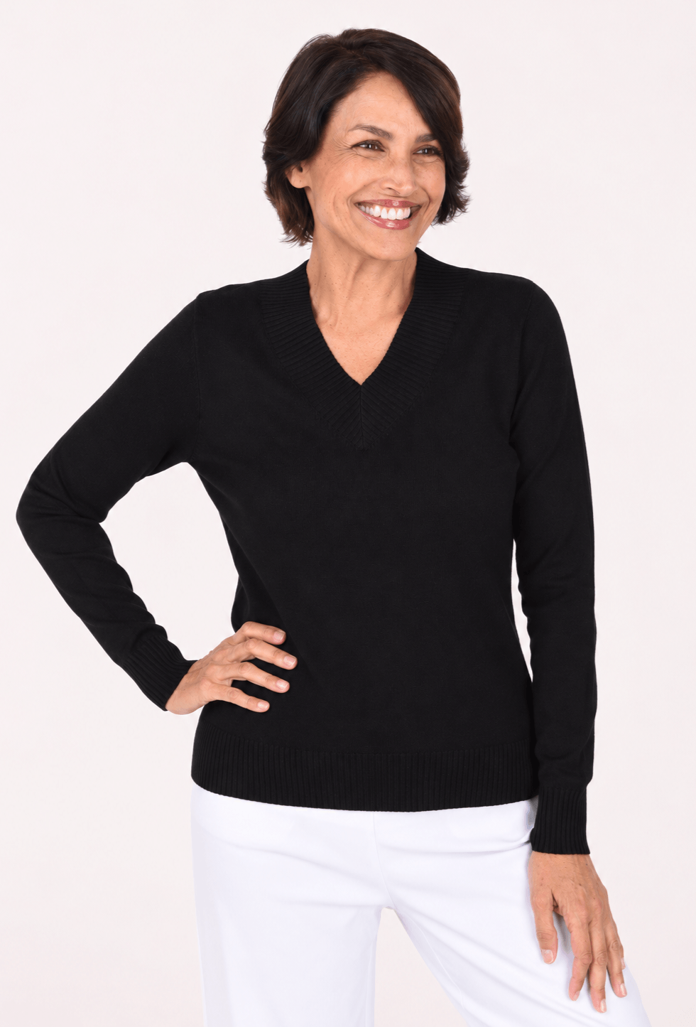 A smiling woman with short dark hair wearing a black long-sleeve V-neck knit sweater and white pants, posing against a plain light background.