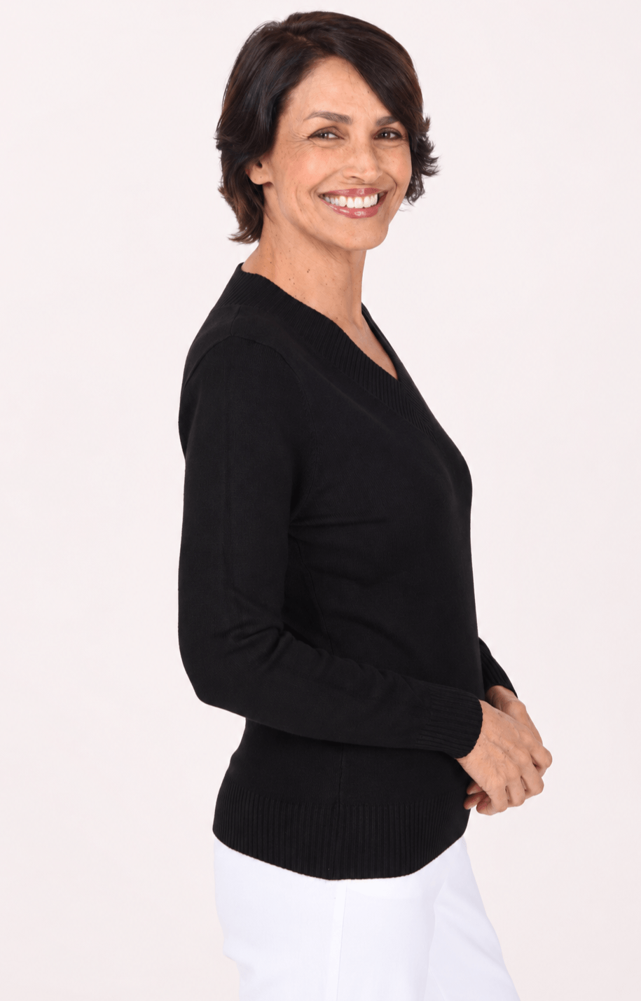 A smiling woman with short dark hair is shown in side profile wearing a black long-sleeve V-neck knit sweater and white pants against a plain light background.