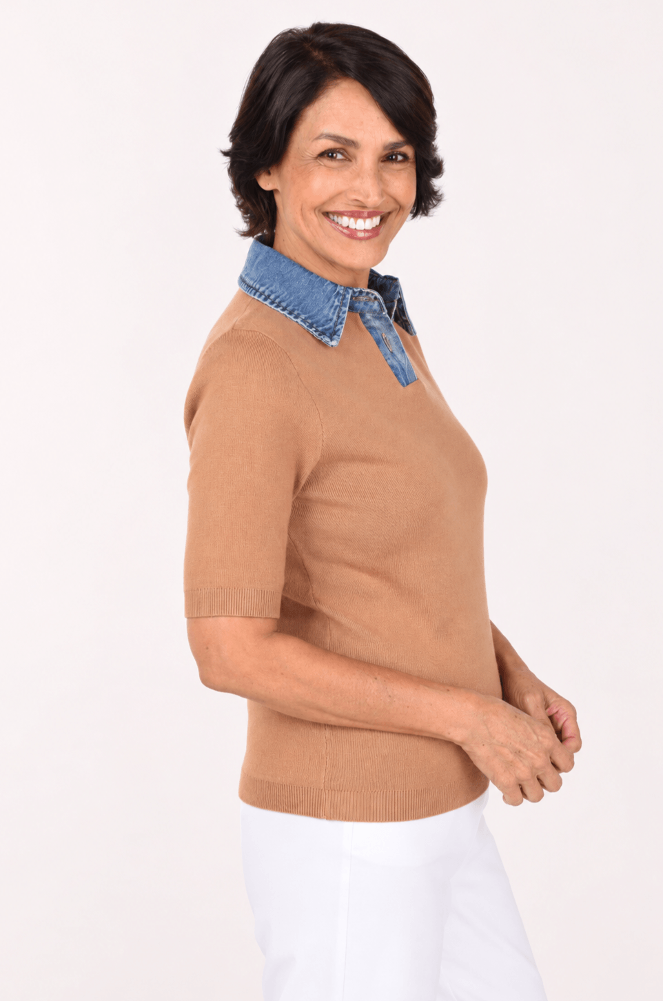 A woman wearing a tan short-sleeve knit top with a blue denim collar, paired with white pants, photographed against a plain light background.