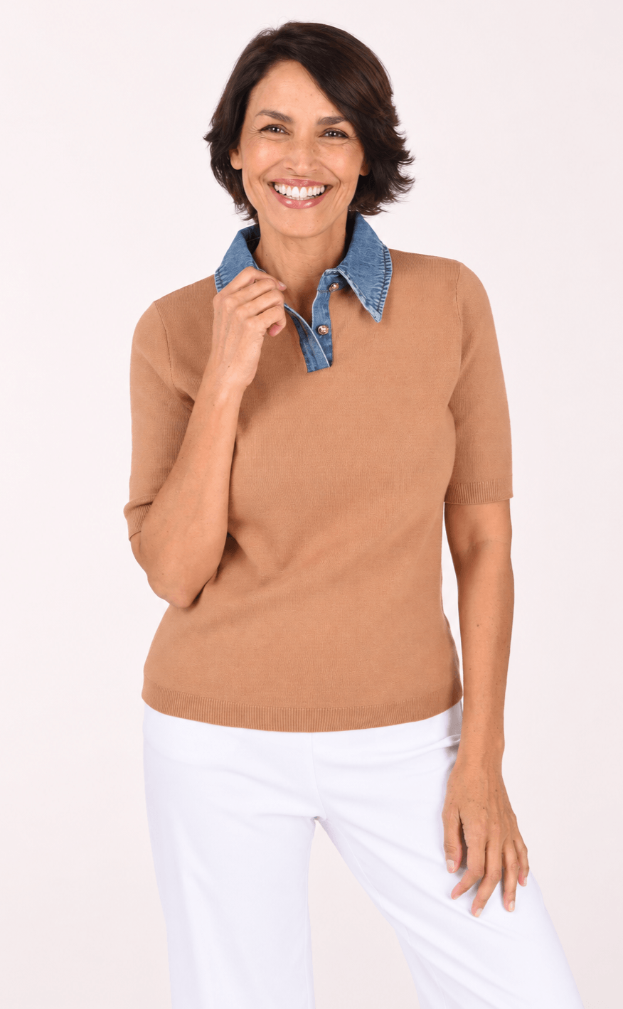 A woman modeling a short-sleeve knit polo shirt in camel brown with a contrasting blue denim collar, paired with white pants, against a plain light background.