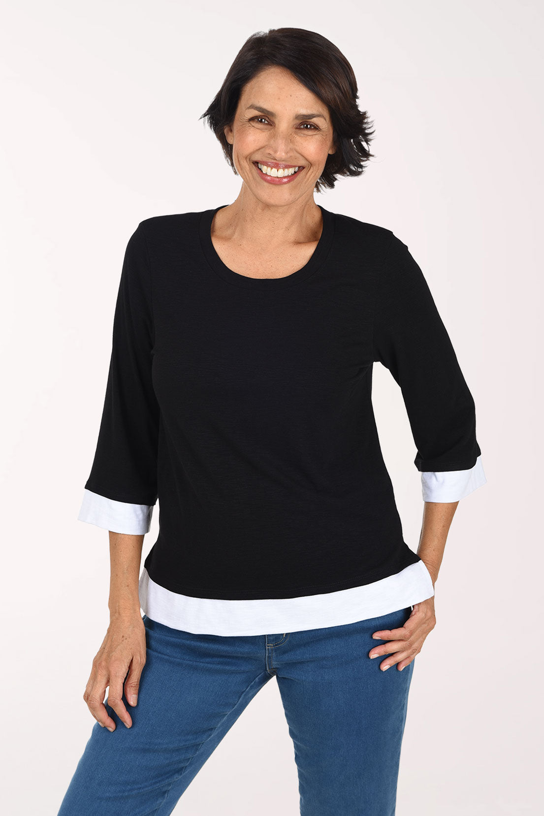 A smiling woman wearing a black three-quarter sleeve top with white contrast trim at the collar, cuffs, and hem, paired with blue jeans, against a plain light background.
