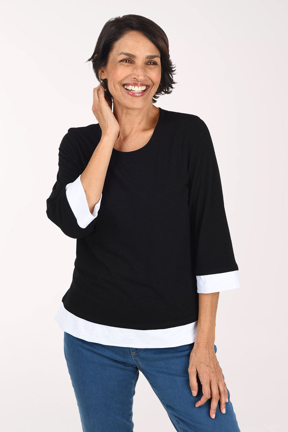 A smiling woman wearing a black three-quarter sleeve top with white contrast trim at the collar, cuffs, and hem, paired with blue jeans, against a plain light background.