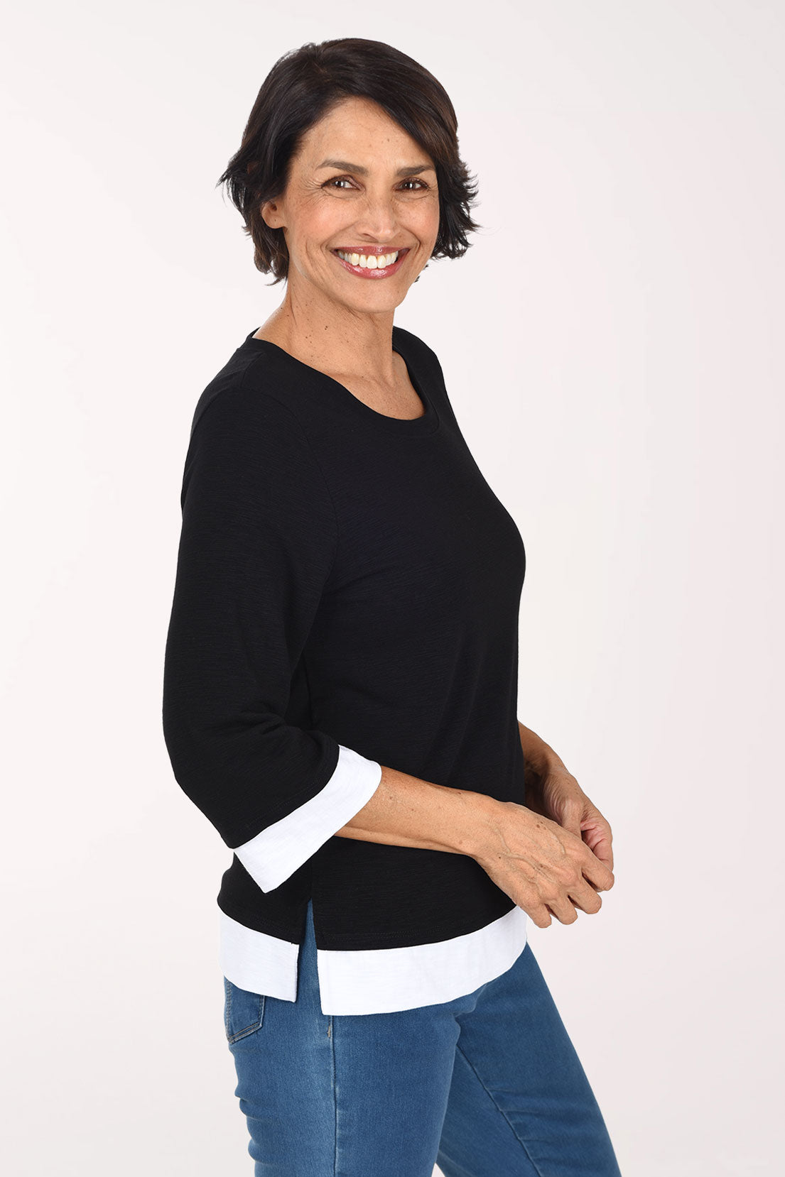 A smiling woman wearing a black three-quarter sleeve top with white contrast trim at the collar, cuffs, and hem, paired with blue jeans, against a plain light background.