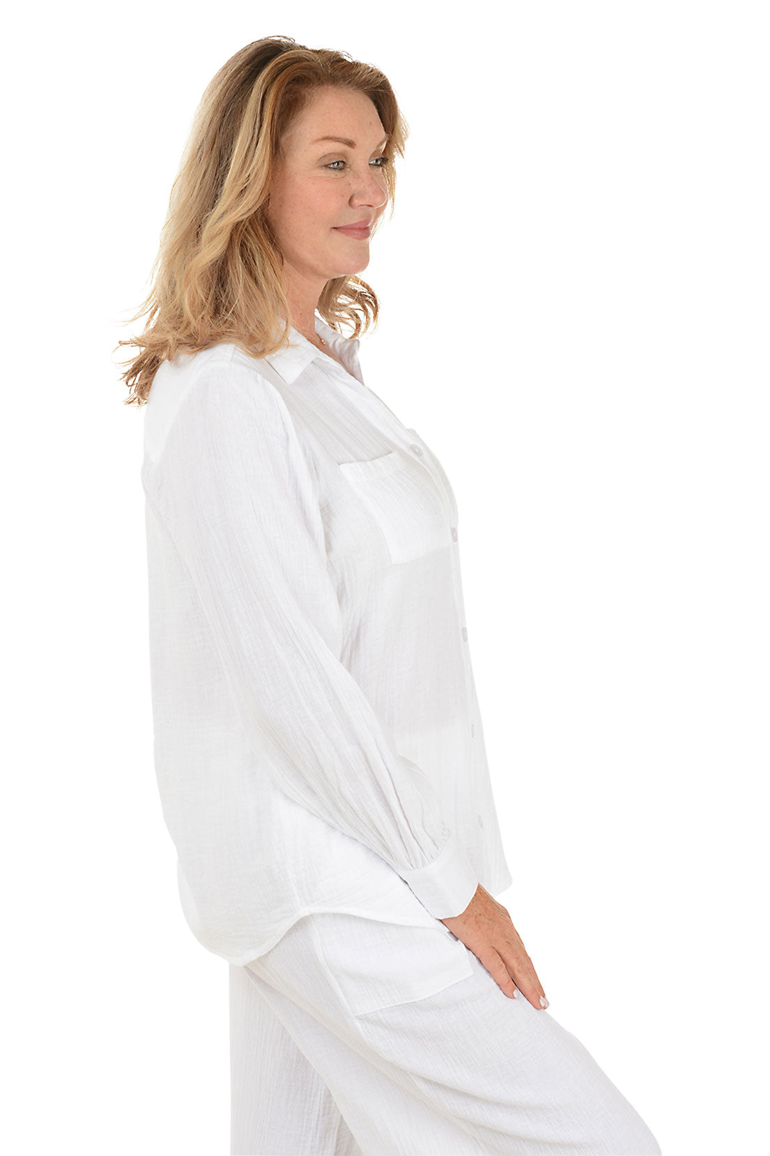 A side shot of a model wearing a white long sleeve cotton gauze shirt with a button-front, foldover collar, double-buttoned cuffs, dual chest pockets, center back pleat, and a curved hemline.