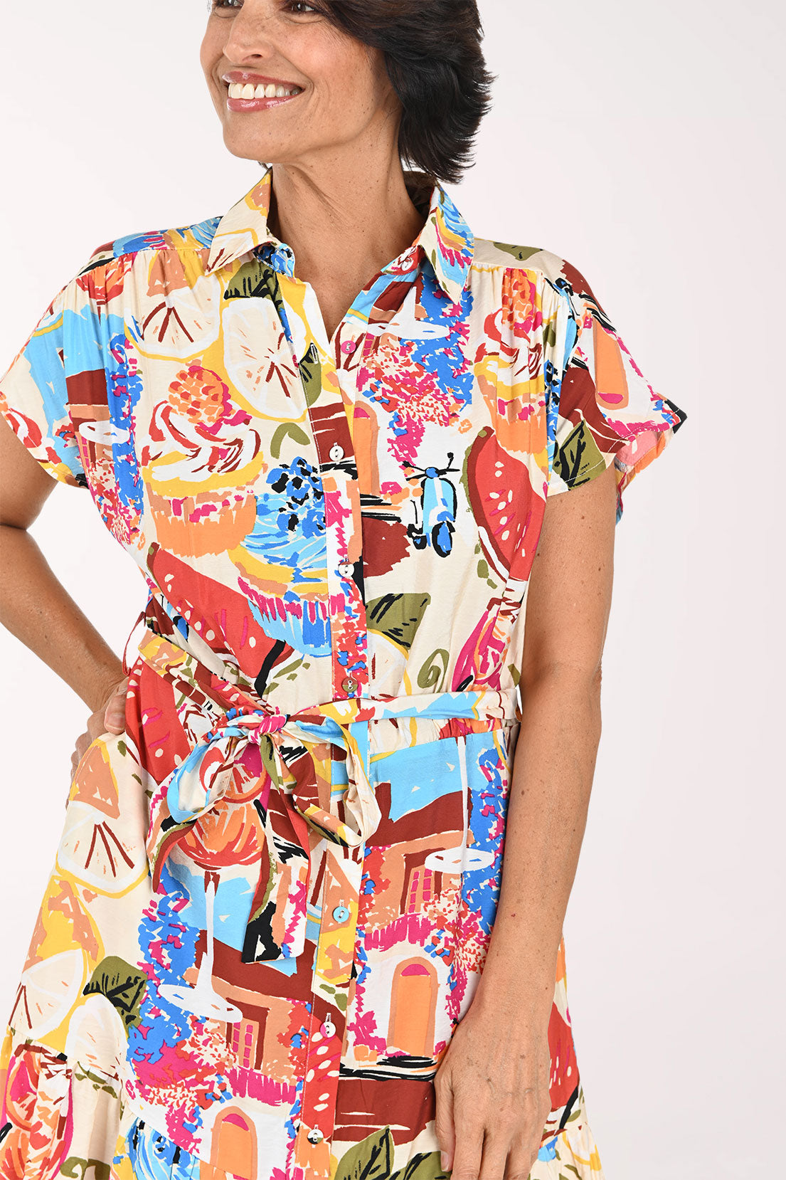 A woman modeling a short-sleeve collared shirt dress with a colorful abstract print, featuring a tie waist and ruffled hem, set against a plain white studio background.