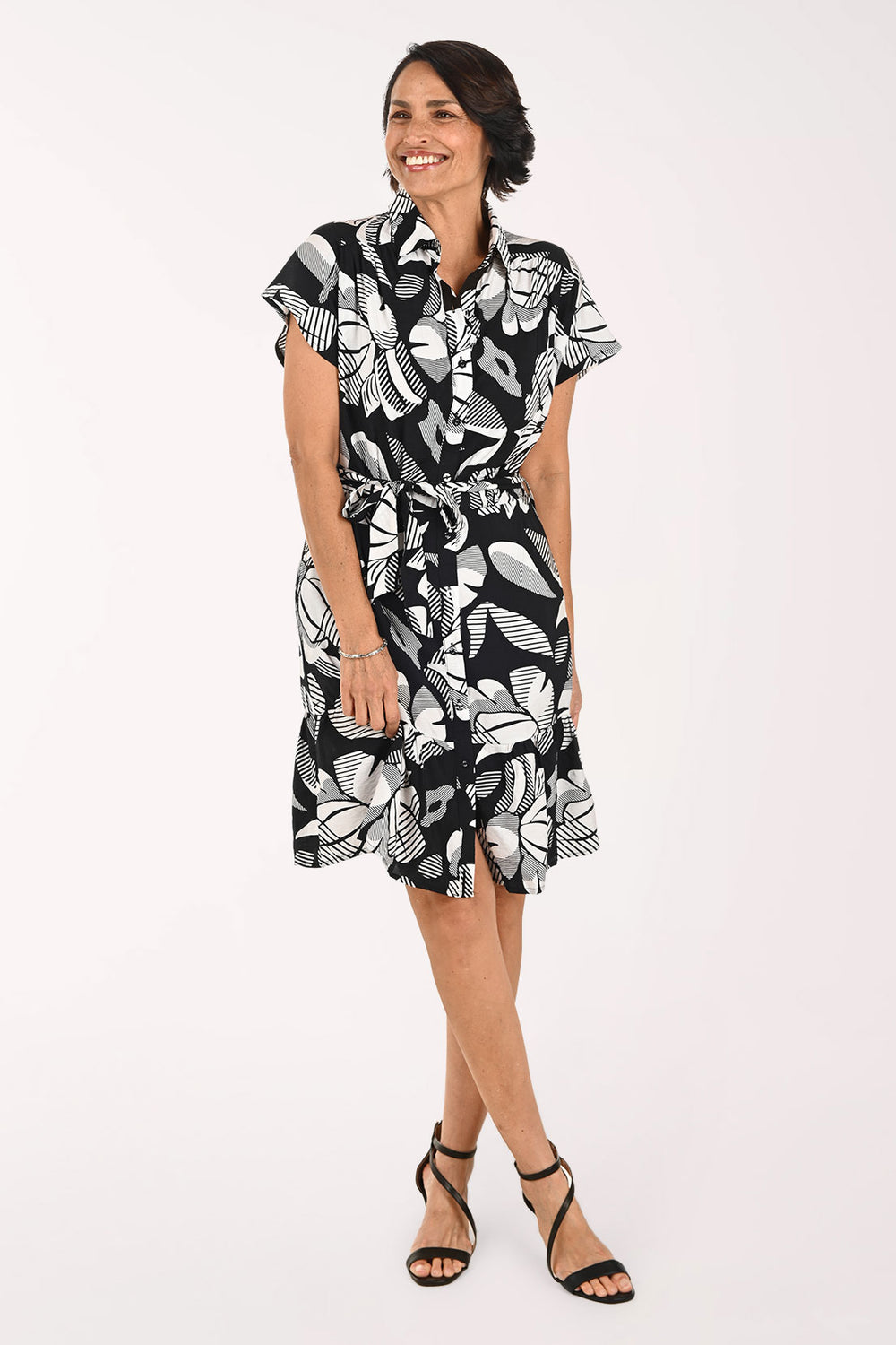 A woman modeling a short-sleeve black and white tropical floral print dress with a tie waist, paired with black strappy high-heeled sandals, against a plain white studio background.