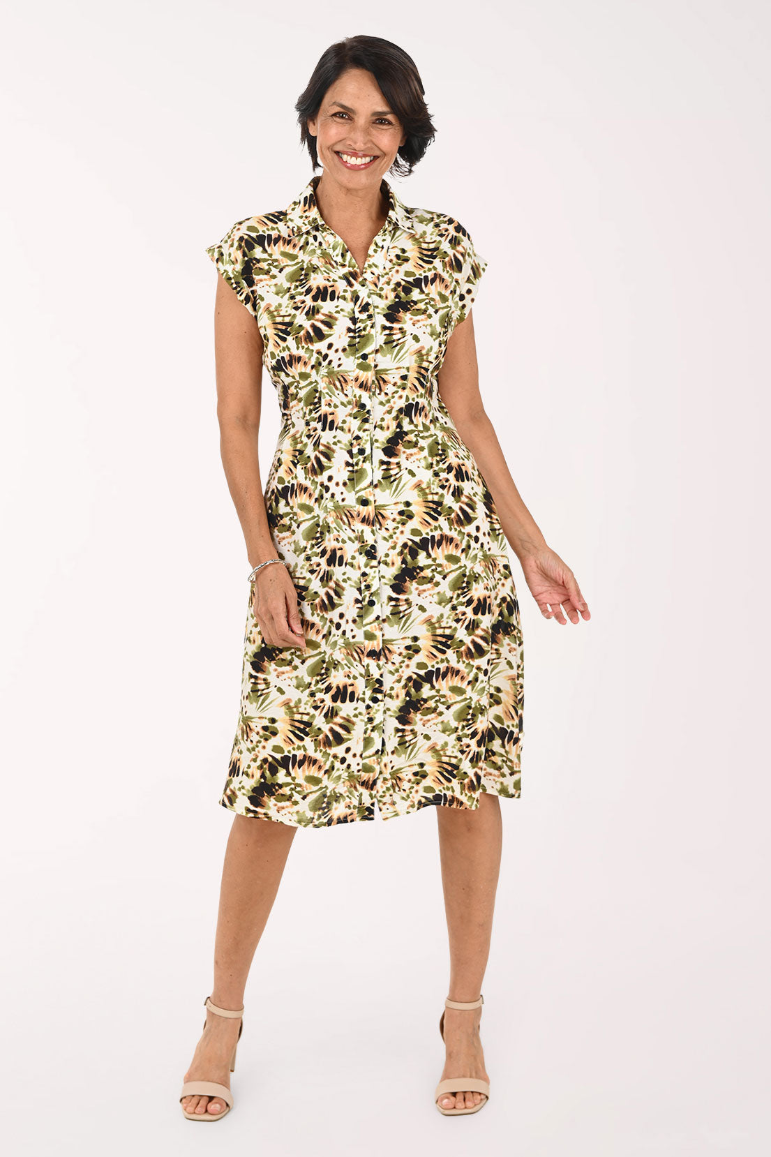 A woman modeling a knee-length floral dress with a cream base and green, black, and brown botanical print, paired with beige heeled sandals against a plain white studio background.