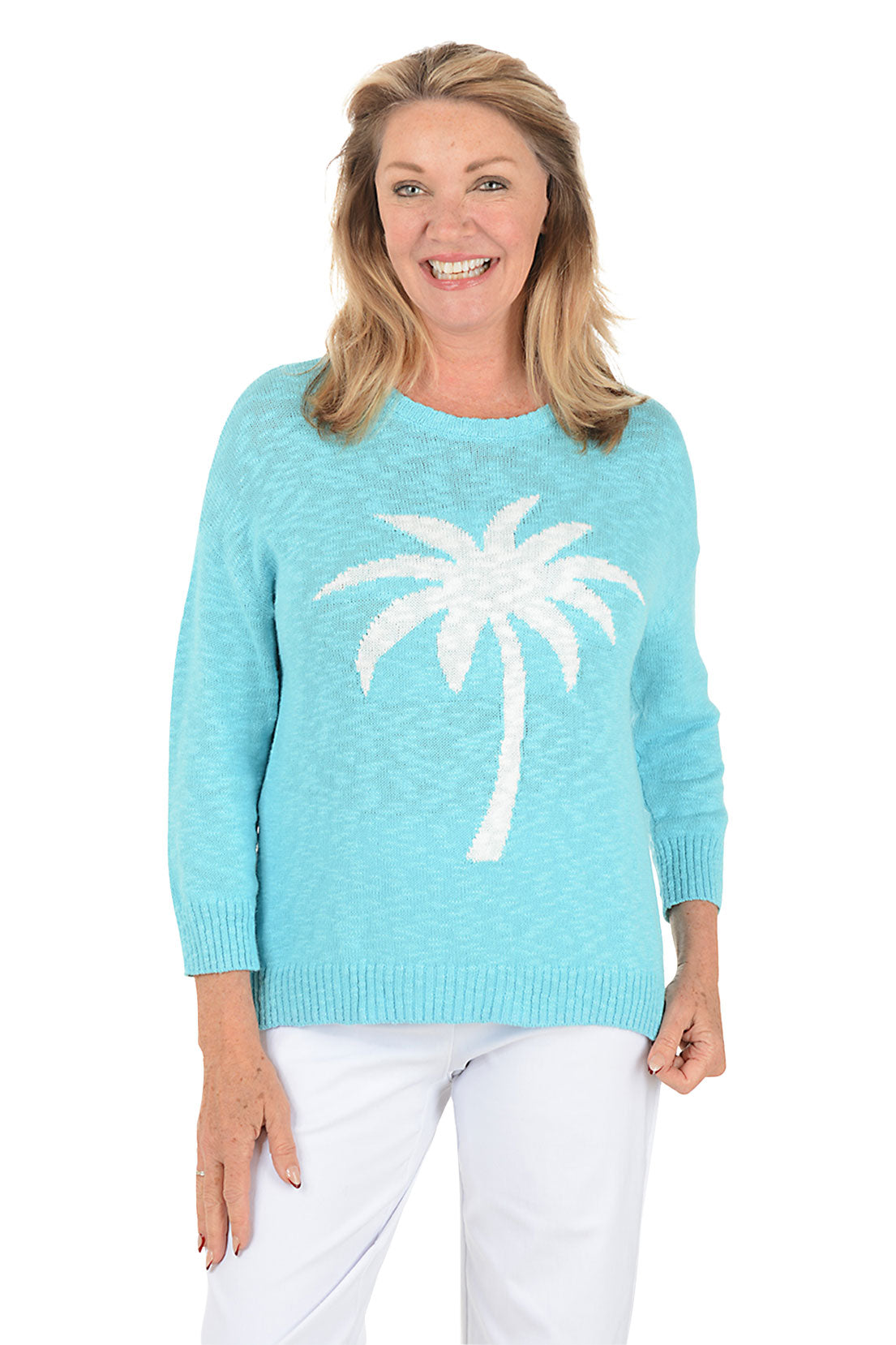 A smiling woman wearing a light aqua knit sweater with a large white palm tree graphic on the front, paired with white pants, against a plain white background.