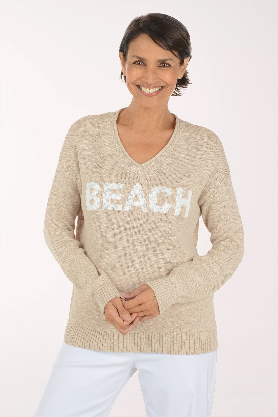 A woman wearing a beige V-neck knit sweater with the word 'BEACH' in large white letters across the chest, paired with white pants, against a plain light background.